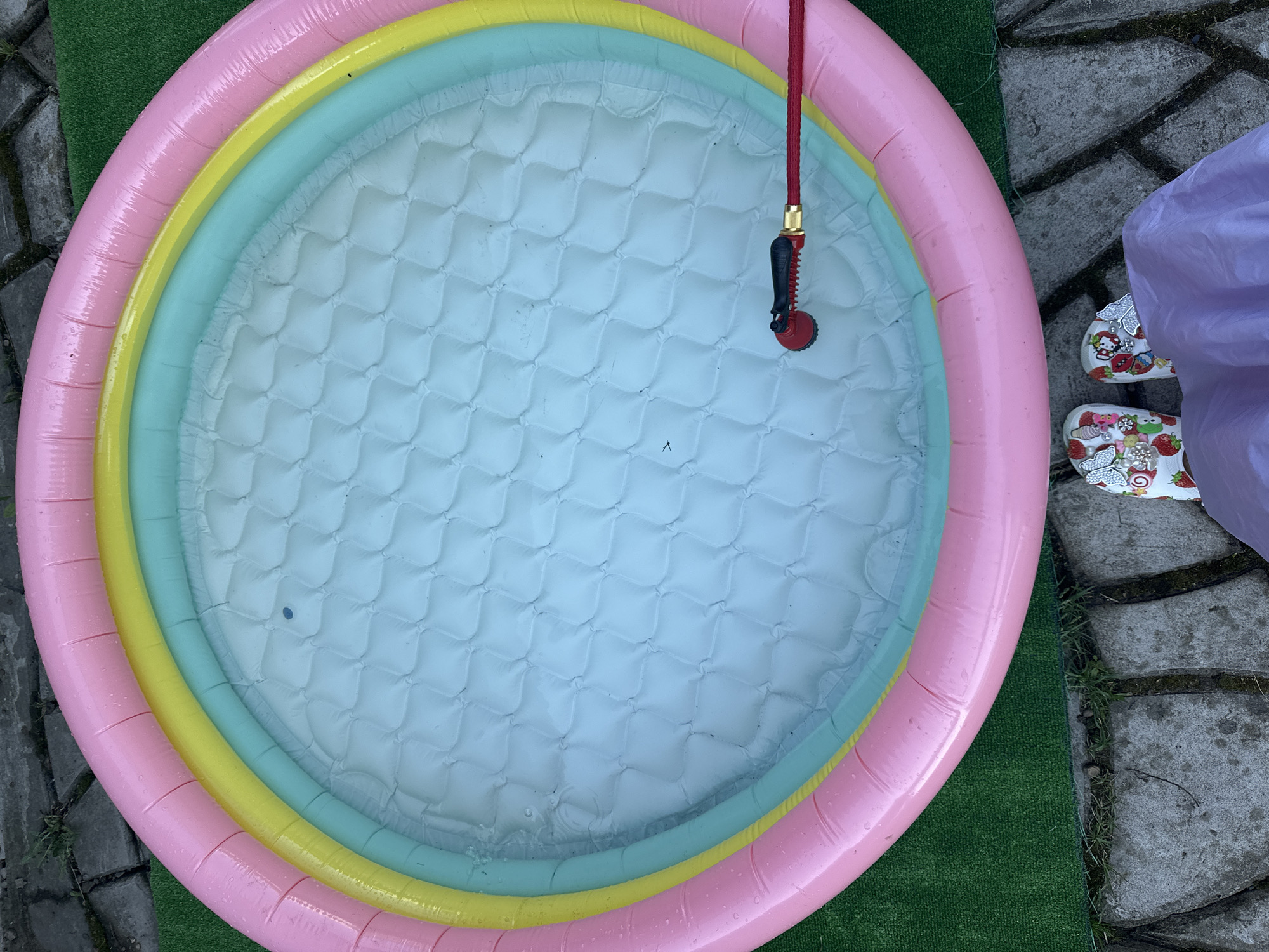 A high-angle shot shows an inflatable children's pool filled with water, sitting on a mixture of artificial turf, brick pavers, and a concrete slab. The pool has three rings of color: pink (outer), yellow (middle), and light blue-green (inner). A red garden hose with a black nozzle is hanging over the edge of the pool, its nozzle submerged. To the right of the pool, near the pavers, are a pair of white sandals with cartoon-themed embellishments and a portion of purple fabric obscuring a person’s leg. The scene is outdoors. The location and city are undetermined, despite the suggestion.