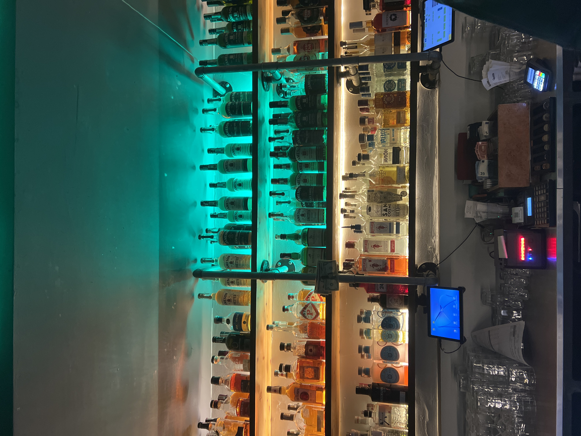 The image shows a well-stocked bar with three shelves of liquor bottles. The shelves are illuminated with warm yellow light, while the wall above the top shelf is lit with a bright green neon light. On the bottom shelf, several rows of clean, empty glasses are stacked. A tablet screen displaying what appears to be a menu or POS system is mounted above the counter, to the left of a receipt printer and a credit card terminal. The overall atmosphere is dimly lit and suggests a commercial establishment, likely a bar or restaurant. The context provided indicates this scene is located in Sofia, Bulgaria. There are no people clearly visible in the image.