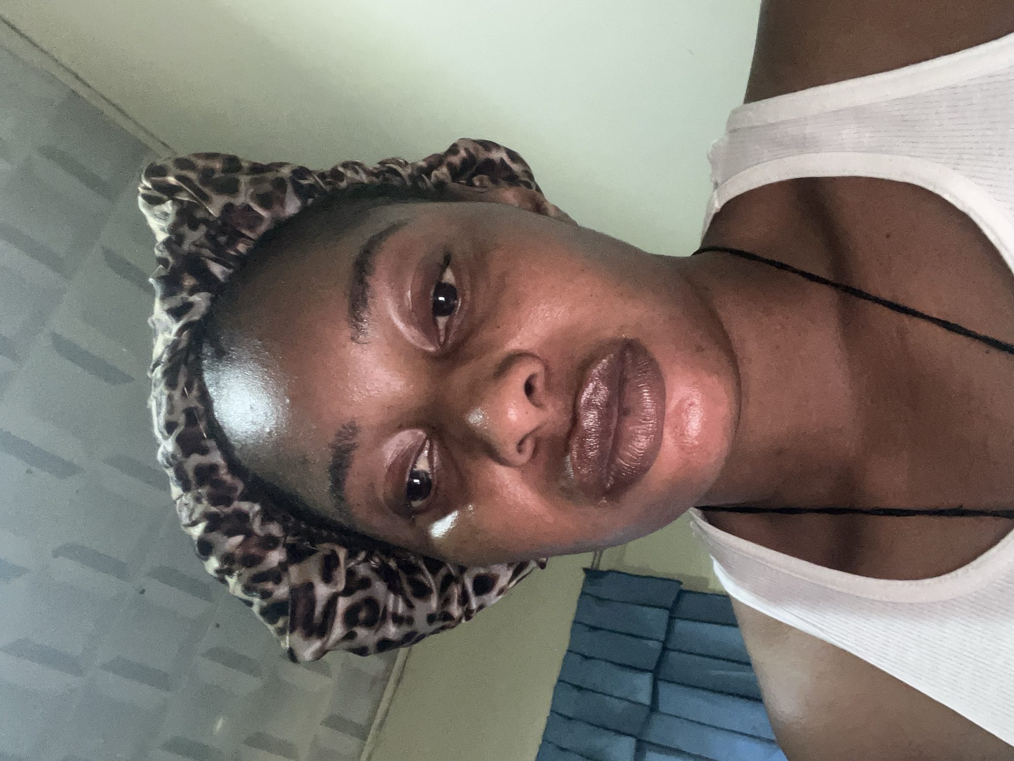 A close-up, low-angle selfie shows a Black woman wearing a leopard print bonnet and a white tank top, looking slightly to her left. She has dark skin and appears to have some light-colored cream applied under her left eye and on her cheek. Her lips are slightly parted and appear to have a dark, glossy lip color. A dark string necklace is visible against her tank top. The background consists of a textured ceiling and a wall with a blue, pleated curtain or blind on the left side. The lighting suggests an indoor setting, possibly a bedroom or bathroom. The overall impression is of a personal moment captured at home.