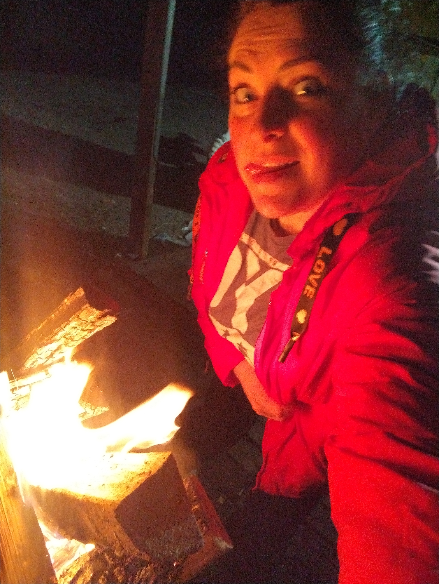 A medium shot taken at night shows a woman posing beside a wood fire, likely outdoors. The woman is fair-skinned with dark hair pulled back. She is wearing a red jacket with the word "LOVE" printed on the drawstring. Underneath the jacket, she's wearing a t-shirt with a white and gray design. The woman has a playful expression, her eyes wide and tongue slightly sticking out. The fire is bright orange and yellow, burning in a fire pit made of wood. The background is dark, suggesting a night-time setting. The photograph appears to be taken at a close range. There is no data to indicate this image was taken in Grishanki, Russia.