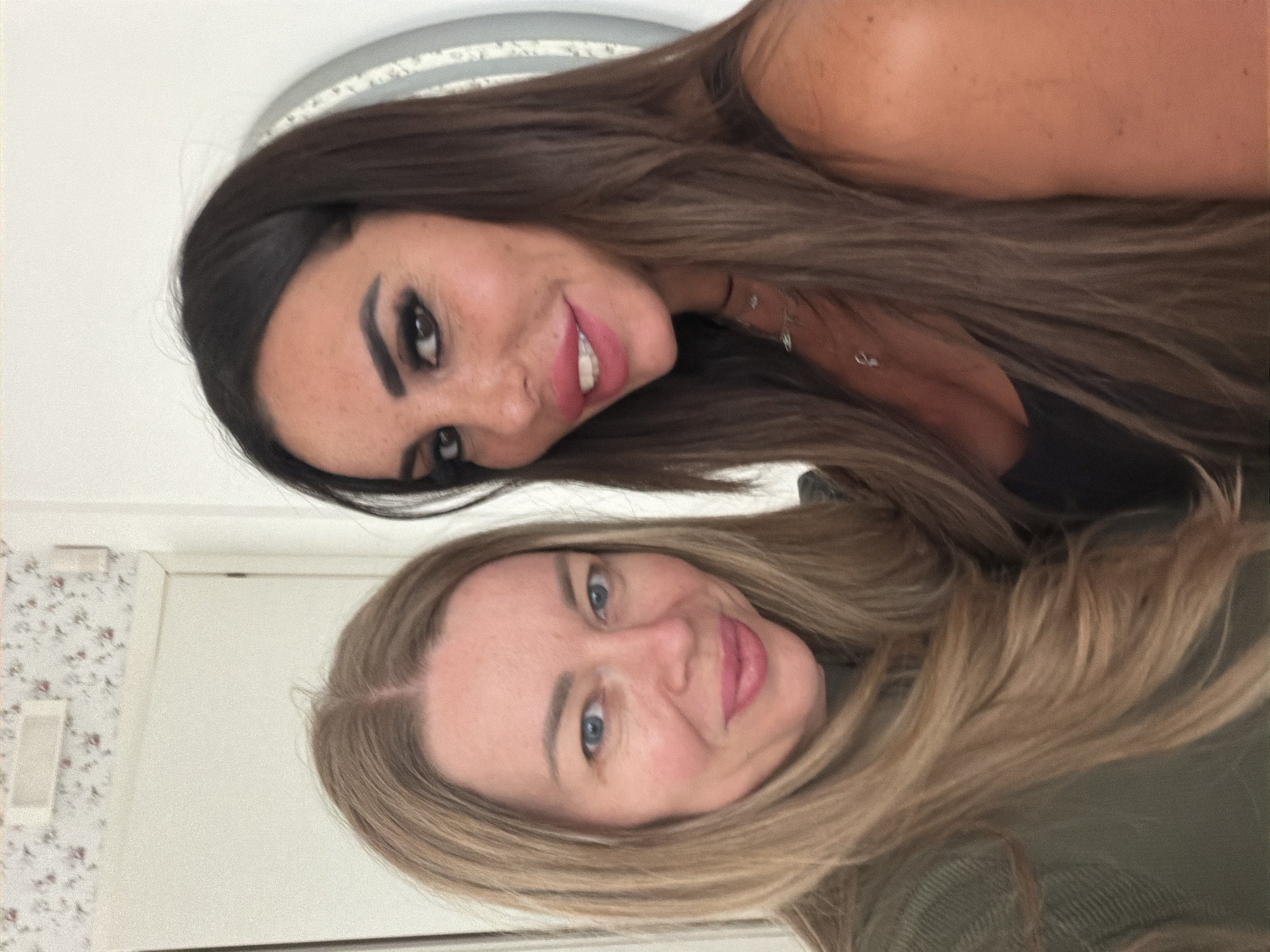 In a selfie, two women are pictured indoors. The woman on the left has blonde hair and blue eyes and is wearing a green top. The woman on the right has dark brown hair, dark eyes, and is wearing a dark top. Both women are smiling. The background appears to be a wall with floral wallpaper and a door. There is no indication that the image was taken in Rome, Italy.