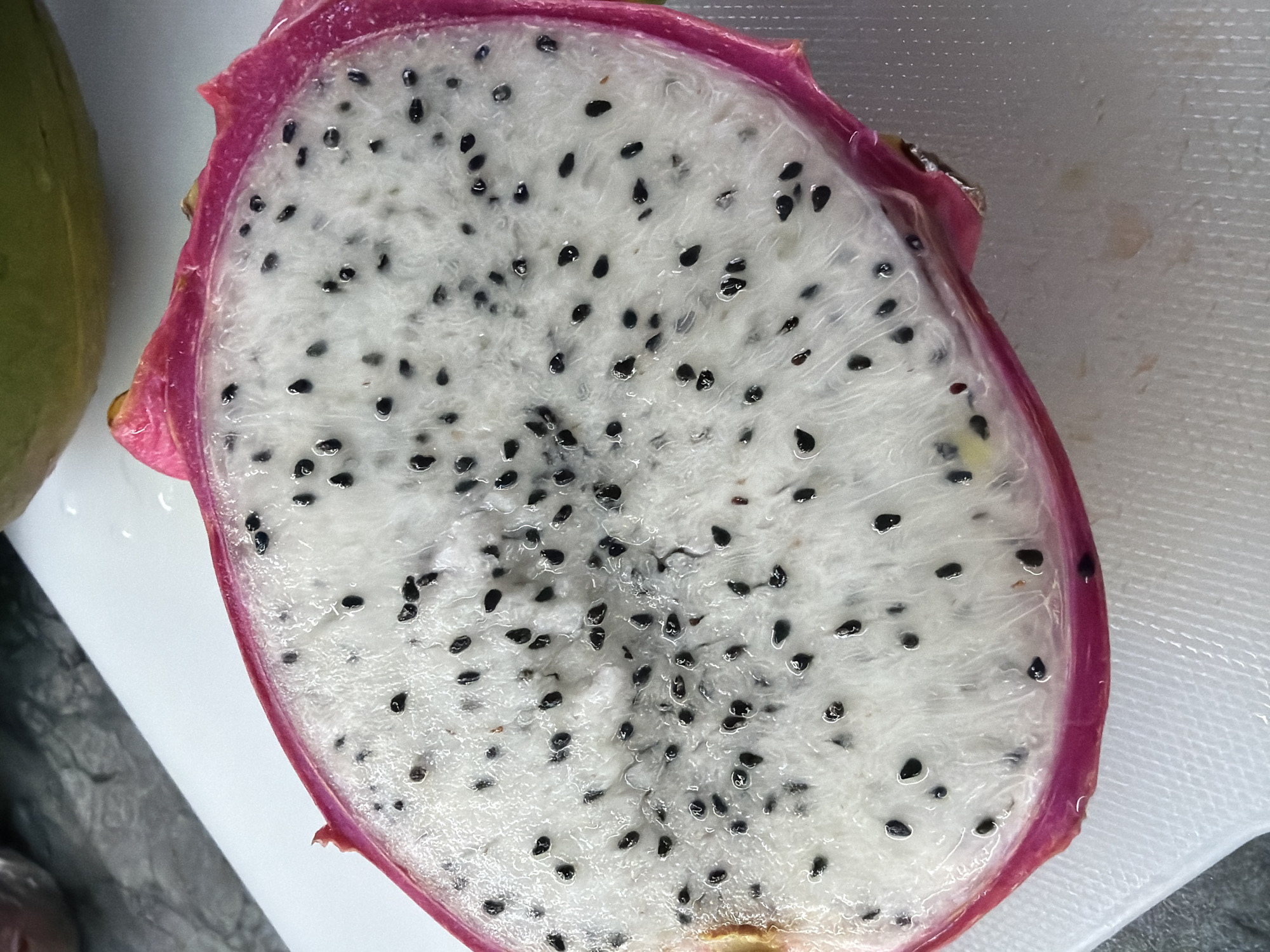 A close-up, overhead shot depicts a cut dragon fruit resting on a white cutting board. The dragon fruit is halved, showcasing its white flesh speckled with numerous small, black seeds. The exterior of the fruit has a bright pink rind with some green patches. To the left of the dragon fruit, a portion of another green fruit is visible. The cutting board sits on a surface that appears to be stainless steel or a similar material. The image appears to have been taken in an indoor setting under bright light. There is no indication of the location being Chaikovsky, Russia.