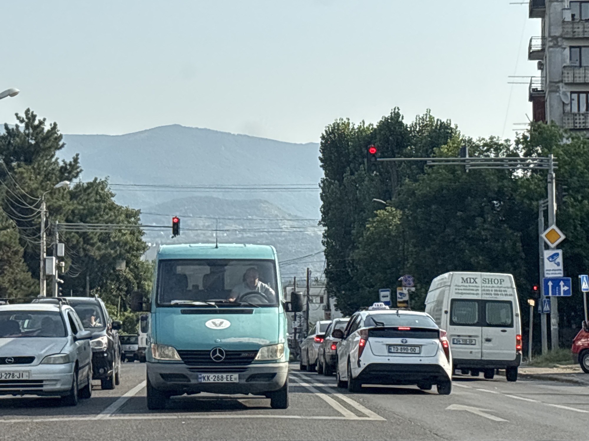 The image depicts a street scene in Tbilisi, Georgia, with a traffic light showing red, indicating that vehicles are stopped. Several cars are lined up at the intersection, waiting for the signal to change. In the foreground, a silver car with license plate "GJ-278-JI" is visible. To its right is a teal-colored van, a Mercedes-Benz Sprinter, with license plate "KK-288-EE". A person is driving the van. Further back, a white van labeled "MIX SHOP" is stopped, and a white Toyota Prius taxi with license plate "TQ-890-00" is also in the queue. The background shows trees, buildings, and distant mountains under a clear sky. Traffic signs and streetlights are present. The overall impression is a typical urban traffic situation during daylight hours.