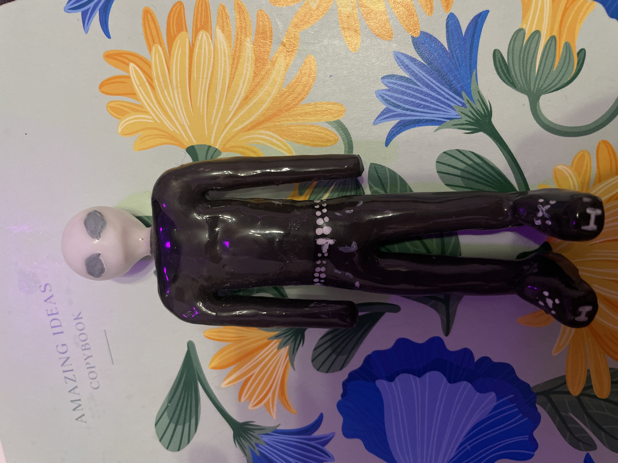 A shiny, dark purple, humanoid figurine with a pale head and grey eyes stands upright in the center of the frame. The figurine appears to be made of clay or a similar material. It is wearing a dark, form-fitting suit with a belt of white circular details around the waist and white accents on its shoes. The figurine is positioned on a surface that has a light blue background adorned with a floral pattern. The flowers are depicted in various colors, including yellow, orange, and blue, with green leaves. Above the figurine, the words "AMAZING IDEAS COPYBOOK" are visible on a lighter blue surface. The lighting in the image casts purple hues on the figurine and the background. There is no indication of the city of Sinyavino, Russia in the image or its context.