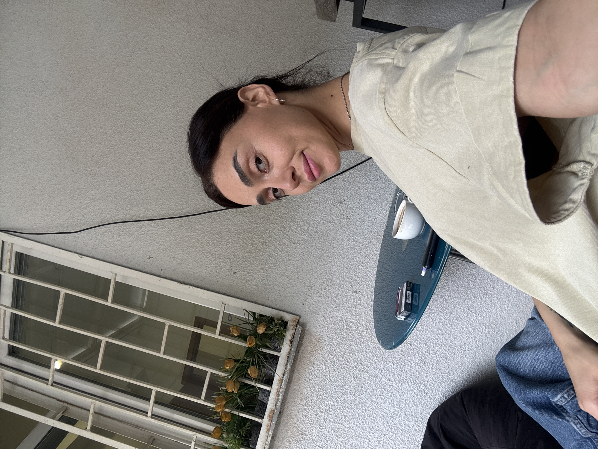 A young woman with dark hair and light skin is sitting at a small blue table outdoors. She is wearing a light-colored t-shirt and blue jeans. On the table, there is a pack of cigarettes, a cup of coffee, and a pen. Behind her is a textured light-colored wall and a window with white bars and flowers in a planter box. The scene appears to be in Tbilisi, Georgia.