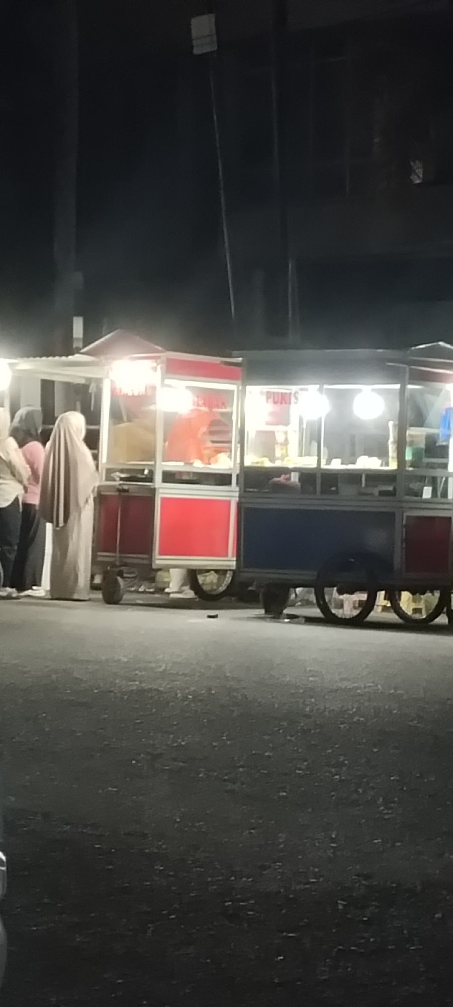 The image depicts a vibrant street food scene at night, likely in Lhokseumawe, Indonesia. Two mobile food carts, constructed with white/silver frames and colorful panels, are prominently featured. The cart on the left has red panels and a small canopy, while the cart on the right features blue and red panels. Both carts are brightly illuminated from within by artificial lights, highlighting the food items displayed inside.
Several people are gathered around the left cart. One individual wearing a light-colored hijab stands facing the vendor, while another person in a darker headscarf and pink top is partially visible behind them. A vendor, wearing an orange top or apron, can be seen working inside the left cart.
The right cart displays the word "PUKIS" in white lettering on a dark sign, indicating that "Pukis," a traditional Indonesian cake, is being sold. The carts are equipped with wheels, suggesting their portability. The foreground shows a dark, textured asphalt street. The background is indistinct and very dark, with faint outlines of poles or structures against the night sky, emphasizing the brightness of the food stalls. The scene captures a typical evening street food environment.