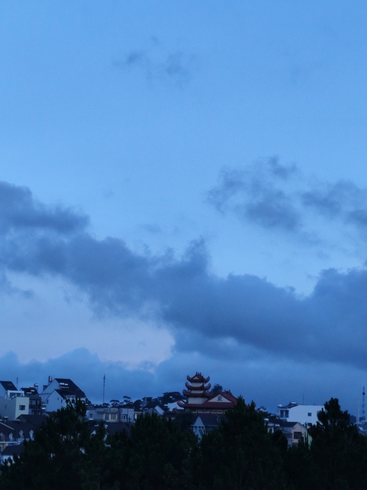 The image shows a nighttime view of a town in Lang Biang - Da Lat, Vietnam, under a cloudy blue sky. A tiered pagoda-style building stands on a hill overlooking the town, which is composed of various low-rise buildings and houses interspersed with dark green trees. The foreground is dominated by the silhouettes of trees.