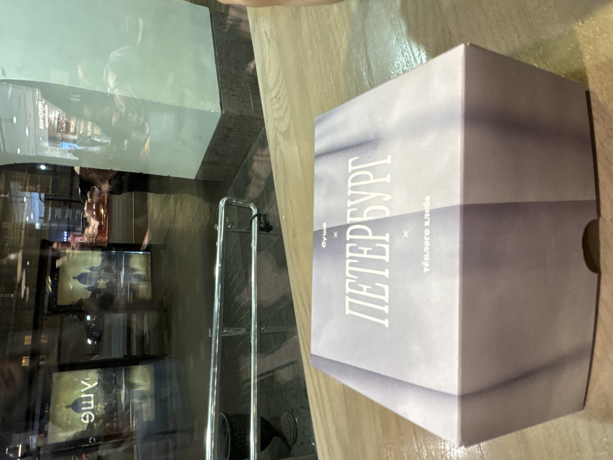 A high-angle shot captures a partially visible takeout box with "ПЕТЕРБУРГ" (Saint Petersburg) prominently displayed in Russian, accompanied by "буше" (Bouche, a restaurant name) and "тёплого хлеба" (warm bread), positioned on a light brown wooden surface. Adjacent to the box is a reflective glass surface with a silver handle. Reflected in the glass are images of Saint Petersburg, including architectural depictions and the word "буше" (Bouche) in reverse. The overall environment seems to be indoors, potentially a café or restaurant setting in Saint Petersburg, Russia.