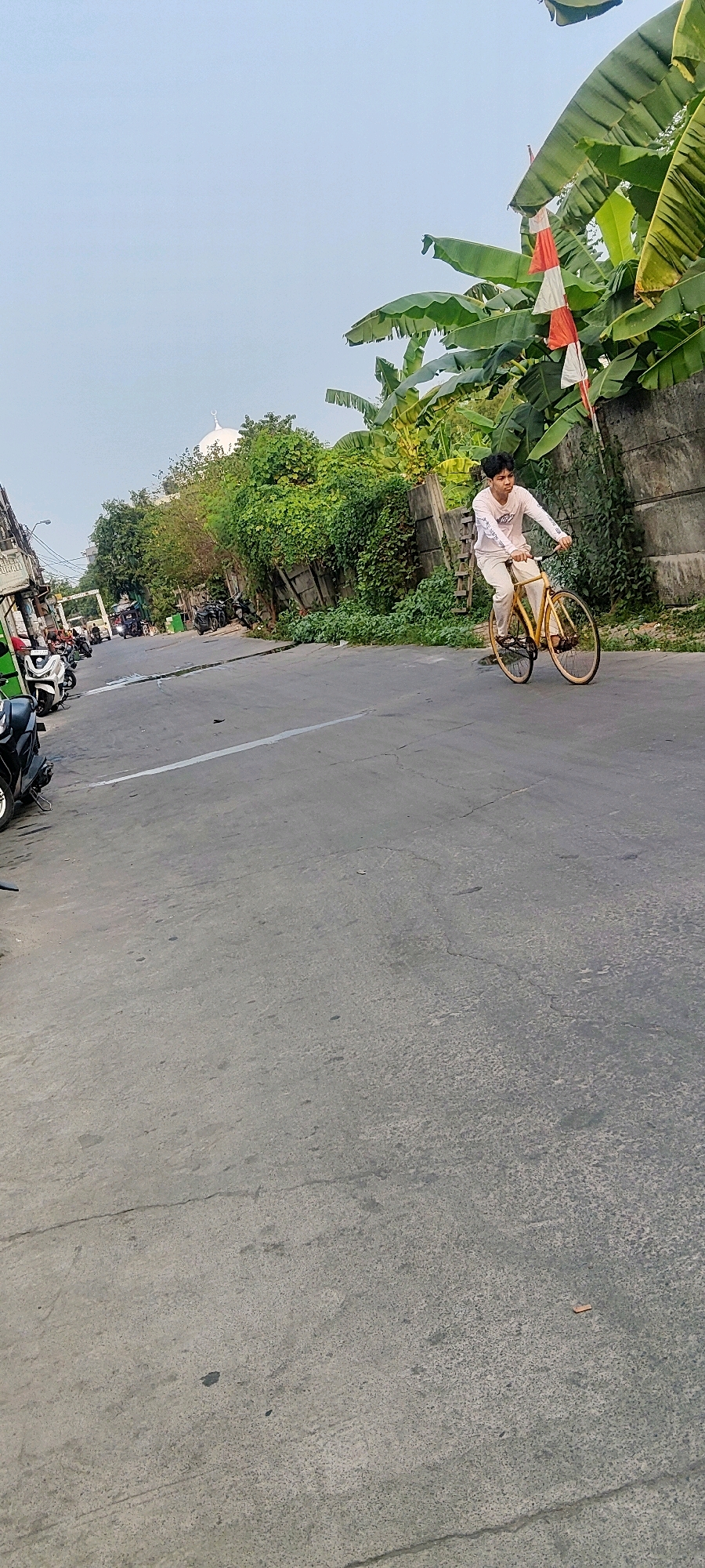 A young person rides a yellow bicycle on a paved road in Mekarmukti, Indonesia. The person, wearing a light-colored long-sleeved shirt and pants, is in motion. The road is bordered by a concrete wall on the right, with lush green foliage and banana trees growing alongside it. A string of red and white triangular flags hangs from a pole amidst the greenery. In the background, on the left side of the image, there are parked motorcycles and a glimpse of a white domed building, possibly a mosque, with trees and vegetation surrounding it. The sky is a clear, pale blue. The road surface is grey and shows some cracks and imperfections.