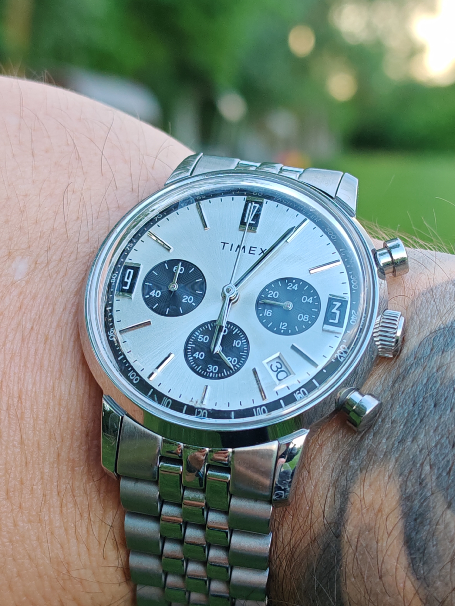 The image shows a close-up view of a silver Timex chronograph watch on a person's wrist. The watch face has a silver background with black sub-dials, numbers, and hands. The date window displays the number "30." The watch has a silver metal bracelet. The person's skin is visible around the watch, with some arm hair and a tattoo. The background is blurred, showing green foliage, suggesting an outdoor setting. There is no indication of the location in the city of Tsarevets, Bulgaria.