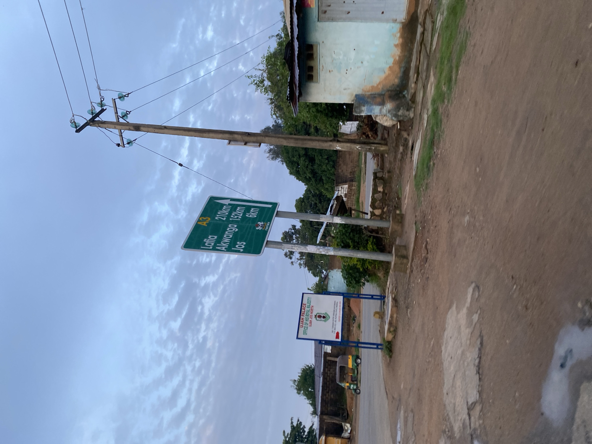 The image depicts a street scene in Jos, Nigeria, under a cloudy sky. A prominent green road sign indicates distances to various cities: Lafia (210 km), Akwanga (152 km), and Jos (6 km). Adjacent to the road sign is a blue and white directional sign for "UJA PALACE OFFICE OF HIS ROYAL MAJESTY UJAH JOS NORTH". In the background, a light blue building with a rusted metal roof is visible, along with trees and a wooden utility pole supporting electrical wires. A three-wheeled vehicle, commonly known as a "keke napep", is parked on the side of the road. The ground is unpaved and appears to be dirt with some puddles and concrete patches. The overall scene suggests a rural or semi-urban setting within the city of Jos.