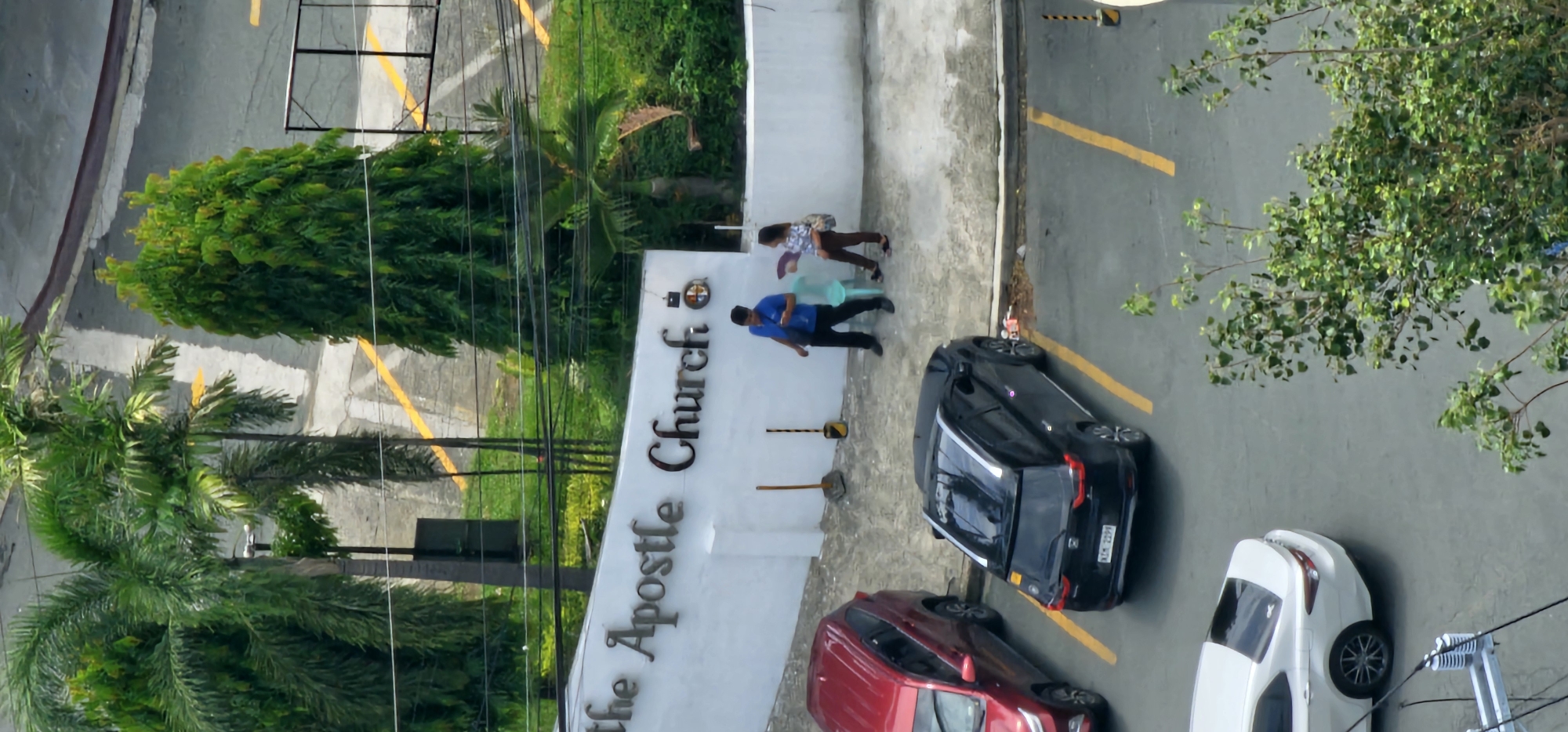 The image shows an aerial view of a church entrance in Makati, Philippines. The church's name, "The Apostle Church," is visible in large metallic letters on a white wall. In front of the wall, two individuals are present: a man in a blue shirt and black pants, and a woman in a patterned blouse and brown pants carrying a bag. A light blue chair is positioned between them. Two cars are parked on the right side of the image: a black SUV and a red SUV. A white sedan is parked on the far left. Trees and foliage are visible in the background, suggesting a tropical environment. The overall scene depicts an outdoor area near a church, likely in a commercial or residential district.
