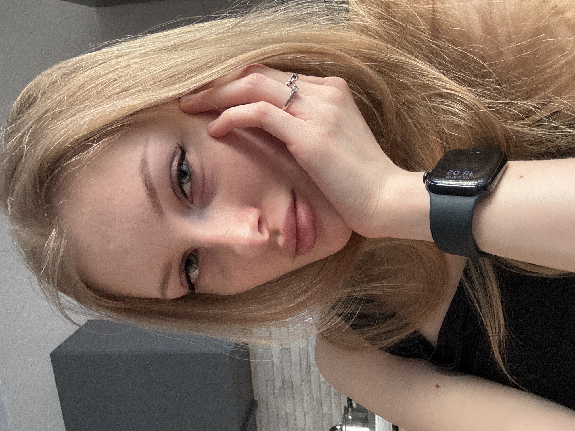 A light-skinned female poses indoors, likely in a personal residence, as suggested by the visible interior elements. Her gaze is directed towards the viewer. She has light blonde hair. Her left hand is positioned near her face, with a ring on her finger. On her left wrist, she wears a smartwatch displaying the time "16:02". The attire appears to be a black top. The environment shows a gray structure in the background, a partially visible light-colored brick wall, and what seems to be part of kitchen equipment. The city of Perm, Russia is not ascertainable from this image.