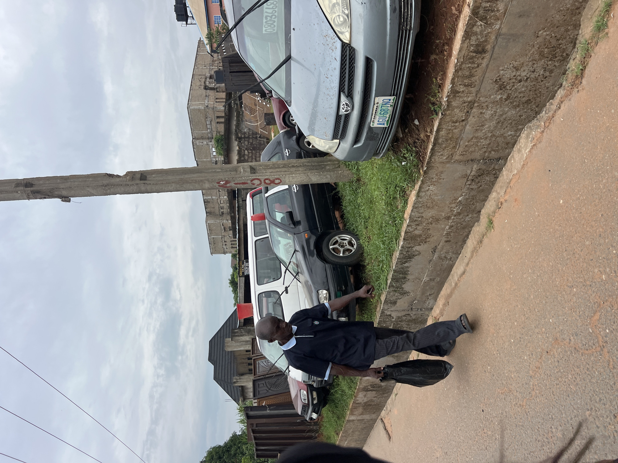 In Nkpor, Nigeria, an elderly Black man walks on a dirt road adjacent to a concrete drainage channel. He carries a black plastic bag in his left hand and wears a dark blue polo shirt with light blue trim on the collar and cuffs, and dark grey trousers. The man walks in front of several parked vehicles, including a red SUV, a white van, a black sedan, and a light blue minivan. A concrete utility pole stands to the right of the man, near the blue minivan. In the background, buildings under construction and other structures are visible. The sky is overcast with grey clouds.