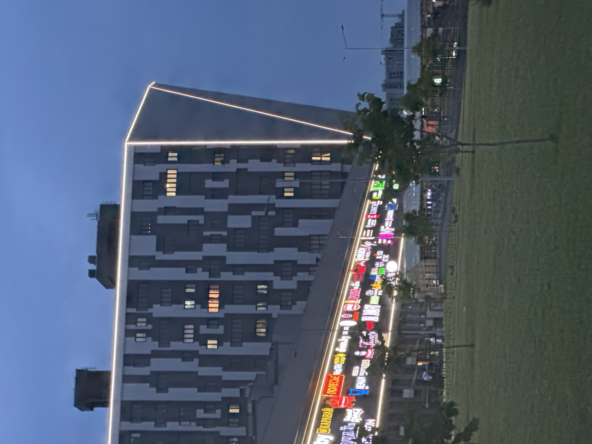 The image shows a modern apartment building with a distinctive patterned facade of grey and white rectangles and illuminated windows. A bright white neon light outlines the upper edge of the building, emphasizing its geometric shape. Below the apartment building, a long, low-rise commercial structure is visible, adorned with a continuous strip of colorful, illuminated signs advertising various businesses. Among the visible signs are "КАРАВАЙ" and "ПОЛЕСЬЕ". The scene is set against a twilight sky, suggesting it is evening. In the foreground, a neatly mown grassy area with a few young trees and a metal fence indicates a public space or park. In the distance, other buildings, some under construction, and a crane are visible. The overall impression is one of urban development and commercial activity in Minsk, Belarus.