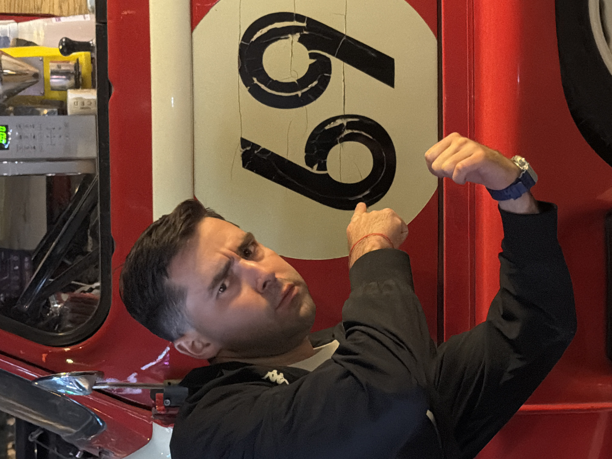 A man with short, dark hair and light skin is captured in a humorous pose, flexing his right bicep and pointing his left hand at a prominent sign. He looks towards the camera with a raised eyebrow and a slight smirk, conveying a playful and confident mood. He wears a black jacket with a white t-shirt underneath, featuring a small white "Kappa" logo on the chest. A blue-strapped wristwatch is on his right wrist, and a red string bracelet is visible on his left wrist.
The man stands in front of a large, circular sign displaying the number "69" in black, which is positioned on a vibrant red background. The black numbers on the cream-colored sign show signs of wear and cracking. The red surface appears to be part of a vehicle or large piece of machinery, possibly a truck or custom car, given its curved body and visible mechanical components.
In the background to the left, industrial-style equipment is visible, including a chrome exhaust pipe, a lever, and a digital display with green numbers. A partially visible white box with the text "ICED" can also be seen. The setting appears to be indoors, possibly a garage, workshop, or a themed establishment in Krasnoyarsk, Russia.