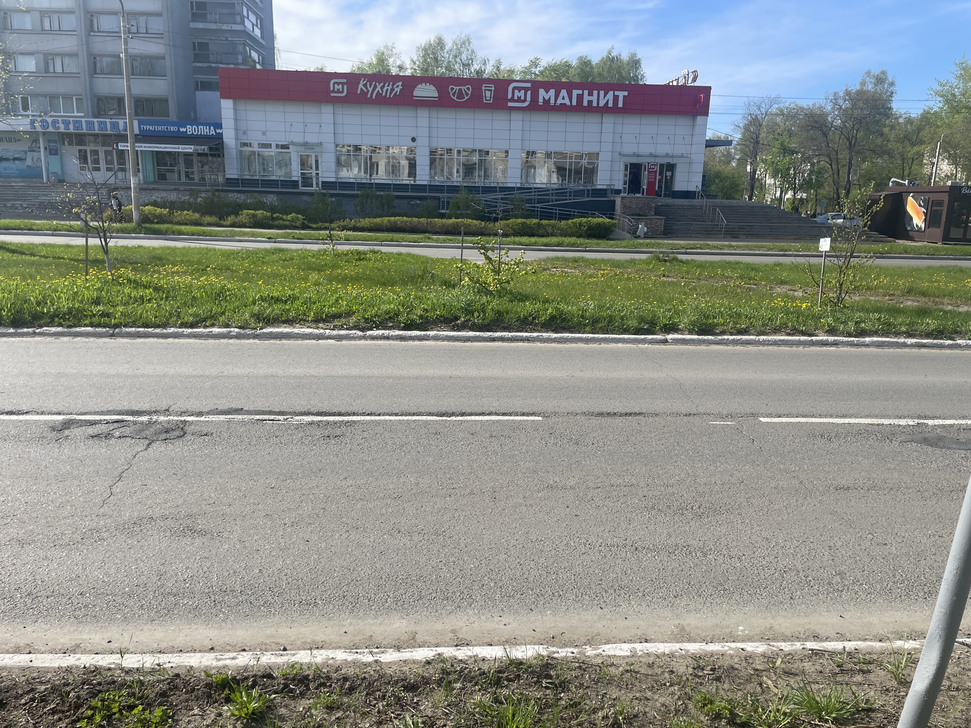 The image depicts a street scene in Chaikovsky, Russia. In the background, a two-story commercial building is prominently featured, identified by a large red sign reading "Кухня" (Kitchen) and "МАГНИТ" (Magnit), indicating it houses both a kitchen supply store and a Magnit supermarket. Adjacent to this building is a multi-story residential building with blue signage indicating it houses a hotel ("ГОСТИНИЦА") and a travel agency ("ТУРАГЕНТСТВО ВОЛНА"). A grassy median separates the buildings from the viewer's perspective, with small trees and dandelions interspersed throughout. The foreground is dominated by a paved road with a visible dividing line, suggesting two-way traffic. The scene appears to be captured on a sunny day with a blue sky and scattered clouds. The overall environment suggests a typical urban setting with a mix of commercial and residential elements.