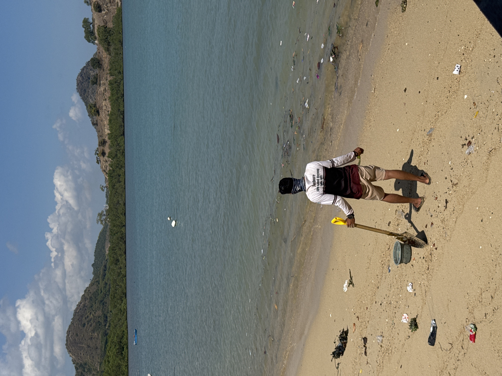 A man stands on a littered beach in Kertasari, Indonesia, holding a shovel and facing the water. The beach is covered with various pieces of trash, including plastic bags, wrappers, and bottles. The water itself is also polluted with debris. In the background, there are hills covered with vegetation and a clear blue sky with scattered clouds. The man is wearing a long-sleeved shirt, shorts, and sandals, and a bandana over his face. A bucket sits on the sand near him. There are a few buoys in the water and a small blue boat in the distance. The scene suggests an effort to clean up the beach, with the man equipped for the task.