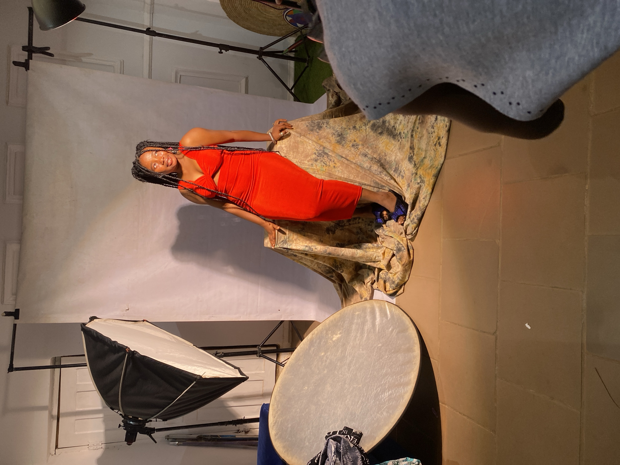The image depicts a studio photoshoot with a Black woman as the subject. She is wearing a form-fitting, bright orange dress and blue, sparkly heels, striking a confident pose while seated on a draped, patterned fabric. Her long braids are a prominent feature. The setting is a studio with a white backdrop, professional lighting equipment including a softbox and a large reflector. The floor appears to be tiled. The lighting suggests an indoor setting, possibly during the day, given the studio environment. The woman's expression is cheerful and engaging. The overall mood is professional and artistic. The provided location context is Jos, Nigeria. There is no visible text in the image.