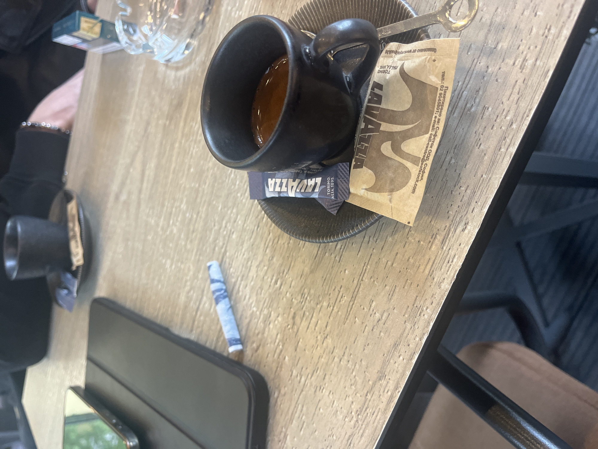 The image presents a close-up, high-angle view of a table in what appears to be a cafe in Plovdiv, Bulgaria. The central subject is a black ceramic espresso cup on a ribbed saucer, containing dark, rich coffee. Adjacent to it are two Lavazza branded sugar packets, one open with a metal spoon resting on it, and the other tucked underneath the cup. The open sugar packet features the "LAVAZZA" logo, "TORINO, ITALIA, 1895.", and Cyrillic text detailing its distribution in Bulgaria: "Пакетирано за: Софсток ООД, София, тел.: 02 9045801; e-mail: call-centre@sofstock.com".
A person's left arm, clad in a black sleeve and adorned with a beaded bracelet, is visible on the left side of the frame. Nearby, a blue cigarette pack and a small, light-colored rolled paper item (possibly a hand-rolled cigarette or filter) are seen on the wooden table. A clear, faceted glass, likely holding water, is also present. Towards the bottom, a smartphone with a reflective screen and a dark case, possibly for a tablet or laptop, are visible. Another small black cup is partially cropped in the bottom left corner.
The scene depicts a casual moment, likely a coffee break or a relaxed gathering. The bright lighting suggests it's daytime. The table has a light wooden surface with visible grain. The presence of coffee, electronic devices, and smoking paraphernalia points to a typical cafe setting.