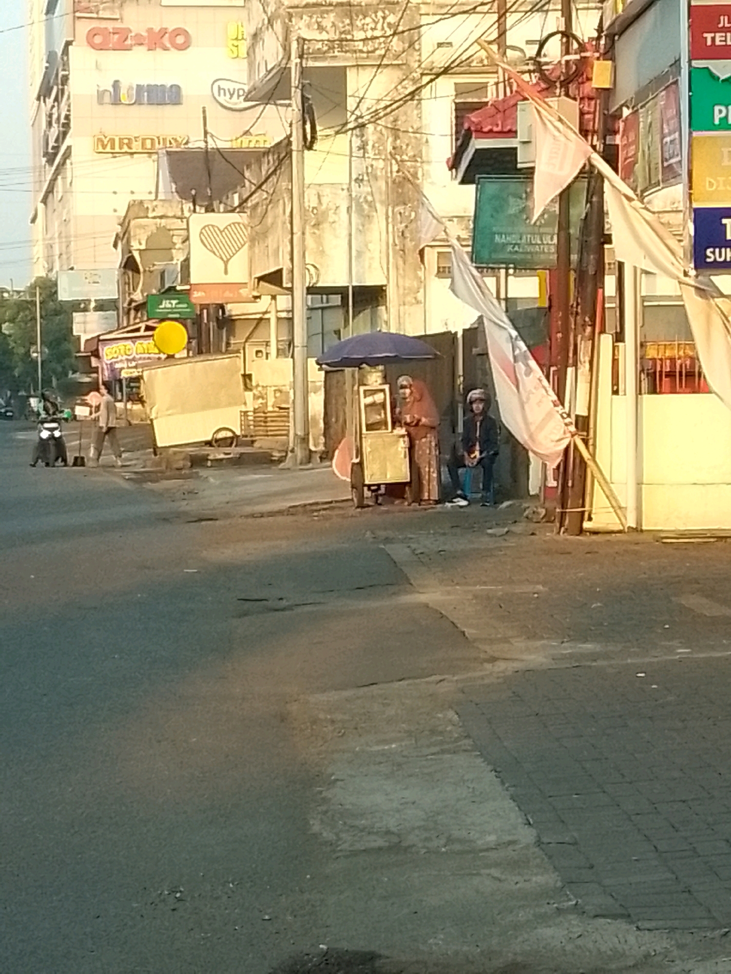 The image depicts a street scene in Jember, Indonesia. In the background, a multi-story building with various advertisements is visible, including "OZ-KO", "informa", and "MR.DIY". To the right, a street vendor with an umbrella and a cart is present, with two individuals sitting nearby. One person is standing next to a motorcycle and holding a broom, suggesting an activity like street cleaning or sweeping. The street itself is paved and appears to be a public thoroughfare. The overall lighting suggests it might be early morning or late afternoon.