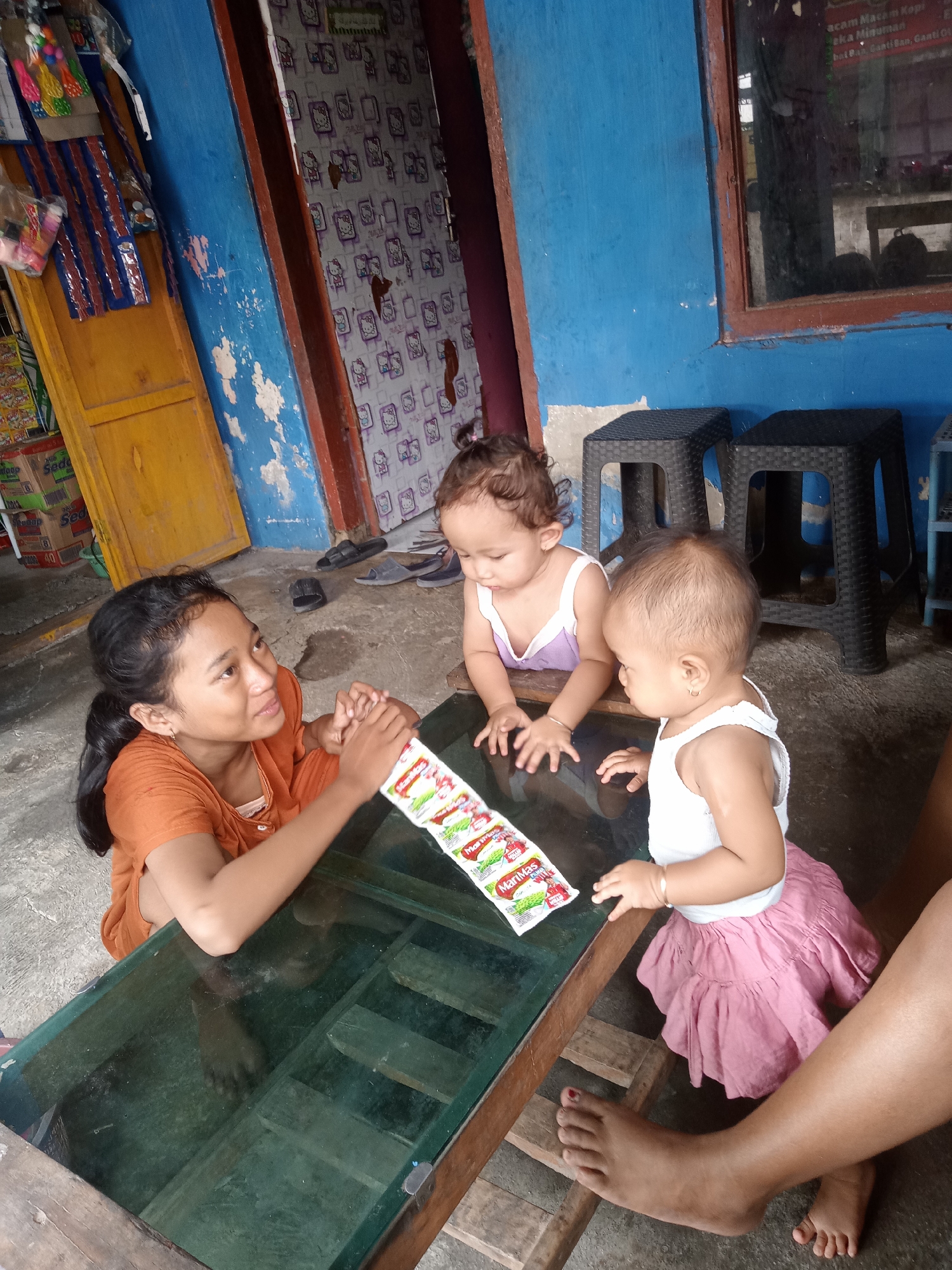 This indoor scene from Bungur, Indonesia, captures a moment between a young woman and two babies gathered around a glass-top table. The young woman, with dark hair tied back, wears an orange top and is leaning on the table, seemingly engaging the infants. She holds a strip of "Marimas" sachets, a powdered drink, which rests on the table.
To her right, a baby with curly hair, in a light purple top, places its hands on the table, looking towards the sachets. Another baby, wearing a white top and a pink skirt, also has its hands on the table, looking to its left. In the foreground, an adult's bare foot with red nail polish is visible, along with a baby's foot, suggesting another person or the woman herself is barefoot and seated nearby.
The setting appears to be a modest home or a small, informal shop. The walls are a striking blue, with noticeable paint peeling. An open yellow wooden door is to the left, and a door with a white and purple Hello Kitty pattern is partially visible behind the young woman. A window with a dark wooden frame is to the right, beneath which sit two black plastic stools.
The bright, natural light streaming into the space indicates it is daytime. Visible text includes "Marimas" on the sachets and "Sedaap" on stacked boxes in the background. A sign near the window displays partial Indonesian text like "acam Macam Kopi" (various kinds of coffee), "eka Minuman" (various drinks), and "bal Ban, Ganti Ban, Ganti Ot" (tire repair/change), hinting at a small business. The interaction appears casual and gentle, with the subjects seemingly absorbed in the moment.