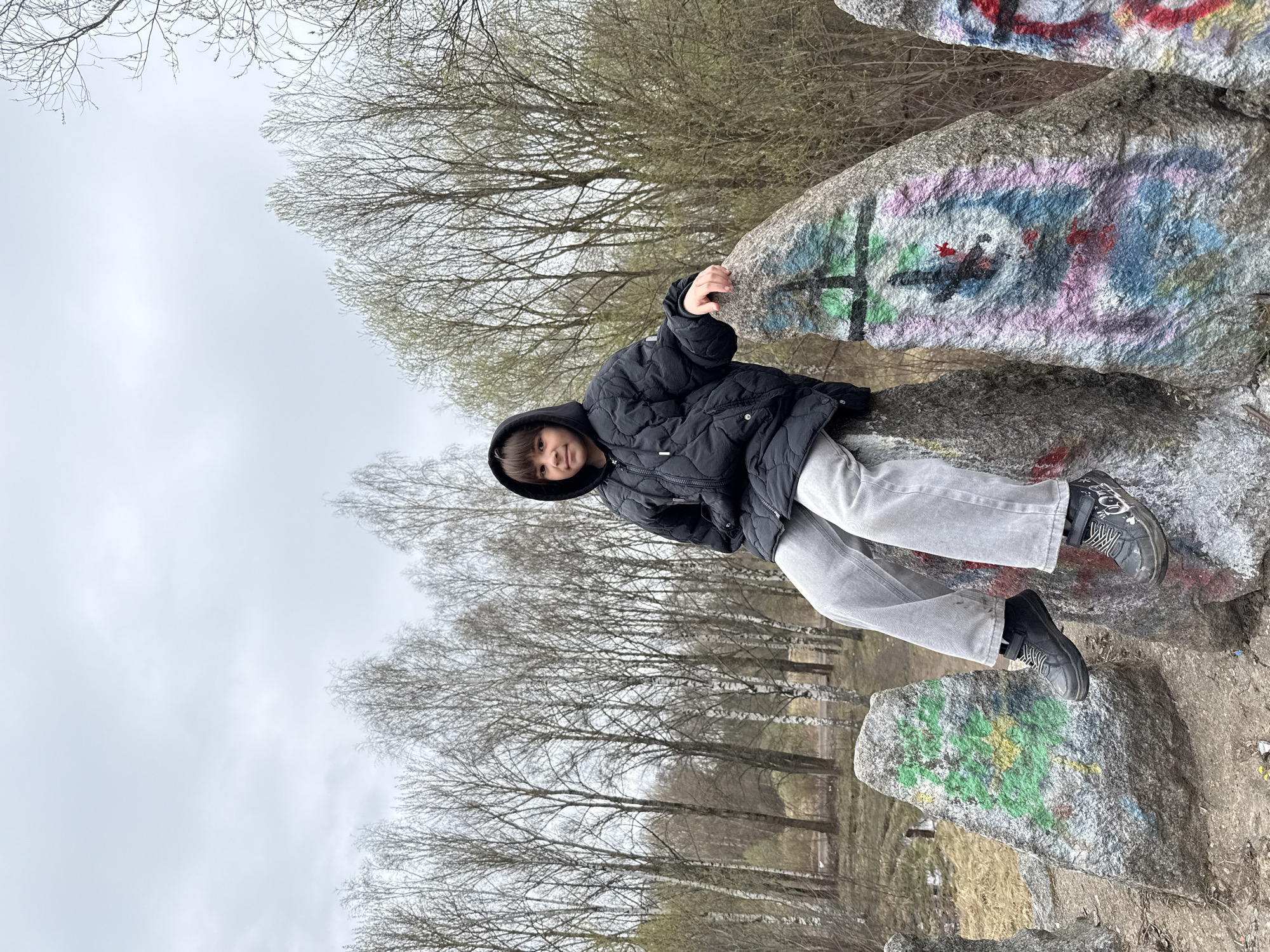 A child wearing a black hooded jacket and light gray pants sits on a gray rock covered in graffiti. The background consists of bare trees against a cloudy sky. The location is Unknown, Unknown.