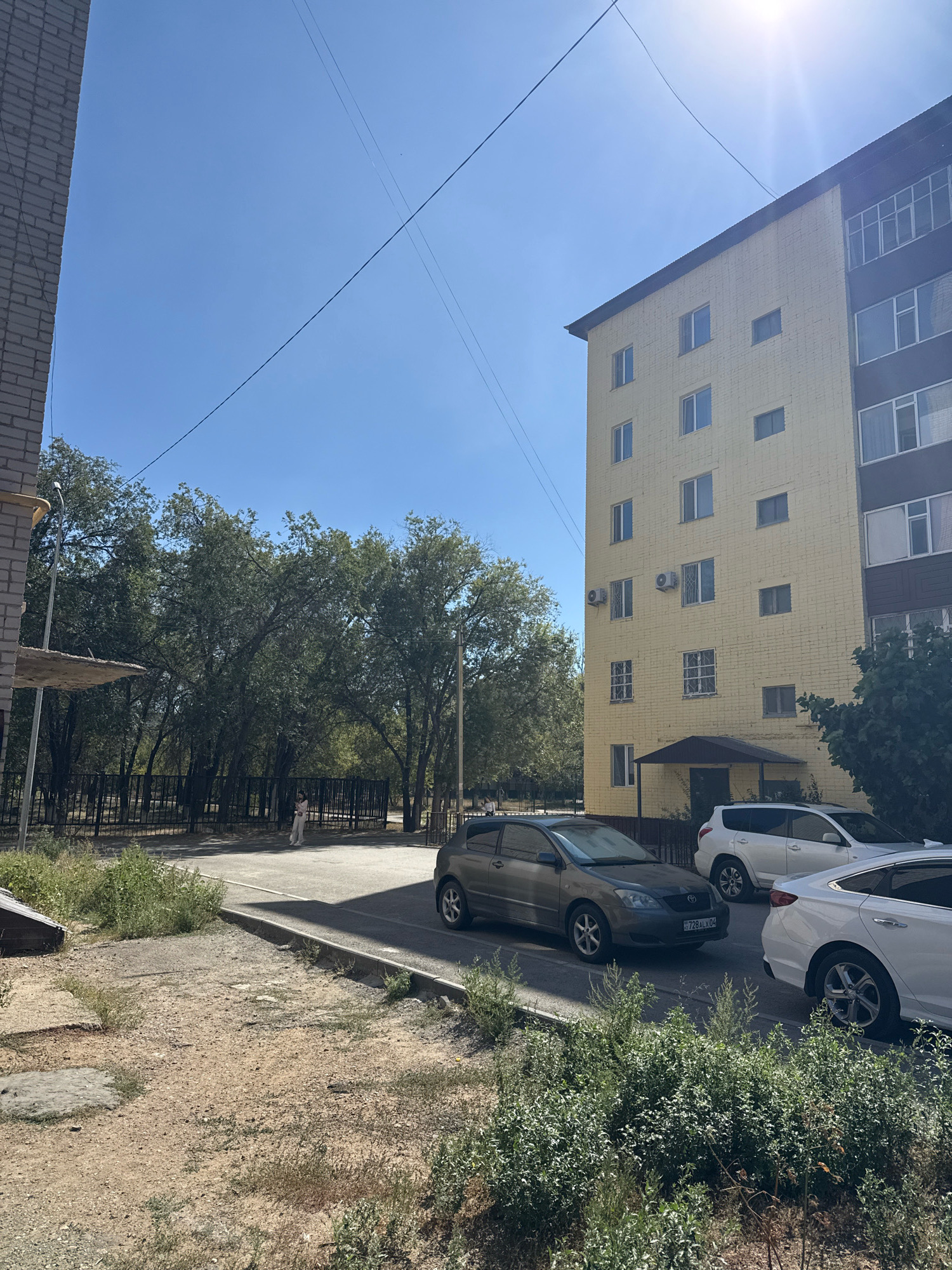 A sunny, clear day in Aktobe, Kazakhstan, with a view of a residential area. In the foreground, dry, patchy grass and dirt with some small bushes. A gray sedan is parked on the asphalt road, facing left, with a license plate visible. Behind it, a white SUV is also parked, facing left. Further down the road, the rear of a white sedan is visible. A black metal fence runs along the side of the road, separating it from a green area with tall trees. A single person in light-colored clothing is walking away from the camera, near the fence. To the right of the trees is a tall, yellow apartment building with many windows. A dark awning is visible above the entrance to the building. The sun is bright in the upper right corner of the image, casting a glare. To the left, a portion of a brick building is visible. Overhead, power lines are strung across the sky.