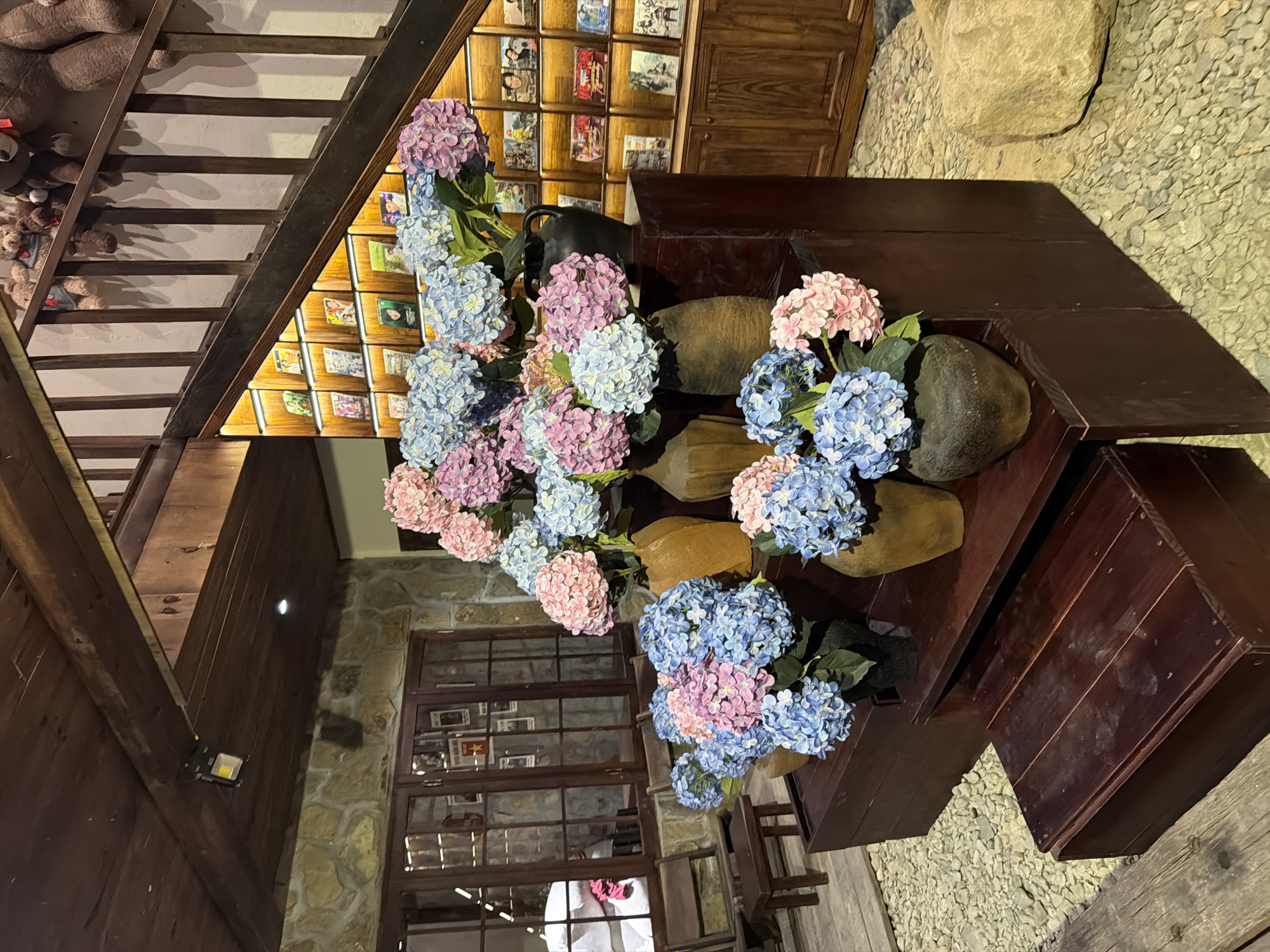 An indoor view within Nam Nha Trang Ward, Vietnam, depicts a rustic space adorned with numerous artificial hydrangeas. The room features a dark wooden plank ceiling with visible beams, one equipped with a modern rectangular LED light fixture. Walls are constructed from irregular light-colored stones. A prominent dark wooden staircase with light risers ascends on the right, displaying multiple brown and beige stuffed teddy bears along its railing and steps. Beneath the staircase, recessed shelves are illuminated and filled with books or small framed items, leading down to a dark wooden cabinet with doors.
In the foreground, a tiered display of dark wooden steps holds multiple arrangements of pink and light blue artificial hydrangeas in various rustic vases, including dark ceramic, tan, and reddish-brown pots. More hydrangeas are visible on the floor and other surfaces. The floor in the foreground is covered with light-colored gravel or small pebbles. On the left, large, dark-framed wooden windows reveal a blurred interior beyond, with posters or images affixed to their glass. Two dark wooden chairs and a small matching stool are positioned near these windows. A large, natural light-colored rock rests on the gravel floor in the lower right. No people are visible in the scene.