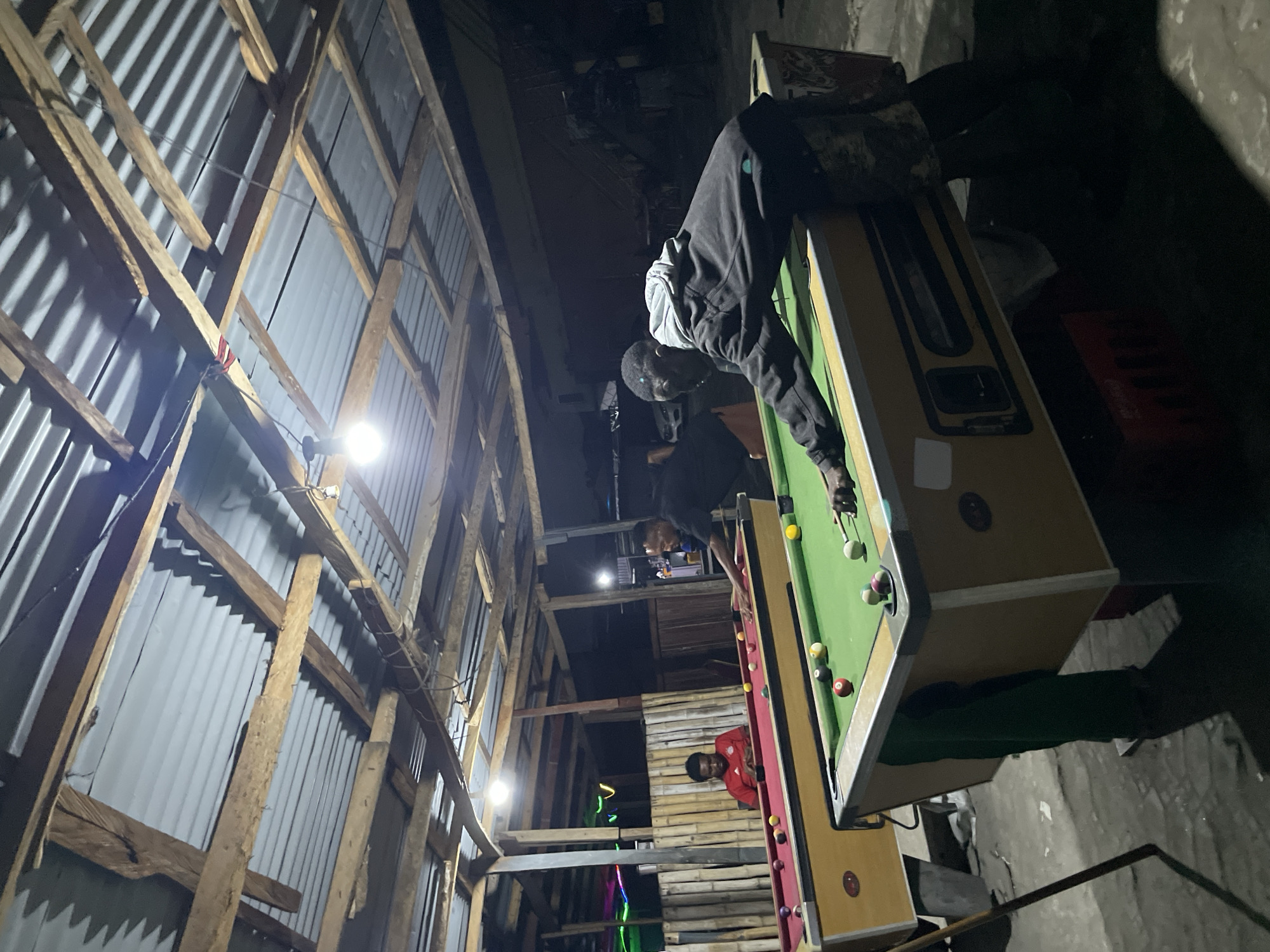 This media file captures an indoor night scene in Ado, Nigeria, within a structure built with corrugated metal sheets and raw wooden beams and planks. The lighting is dim and artificial, provided by bare light bulbs or LEDs casting sharp shadows.
The main activity centers around a game of pool (billiards). In the foreground, a man wearing a dark hoodie with light grey sleeves is leaning over a green-felted pool table, cue in hand, focused on making a shot. Several pool balls are scattered on the table surface. A second pool table, partially visible further back, appears darker in color. Another person in a red shirt is seen in the background, observing the game from a lower level. Additional figures are dimly visible in the shadows.
The setting suggests an informal or utilitarian space, possibly a community recreation center or a local bar. Exposed electrical wiring is visible along the wooden supports. The floor is dark and uneven. The overall atmosphere indicates a social gathering for leisure. No discernible text is visible in the image.