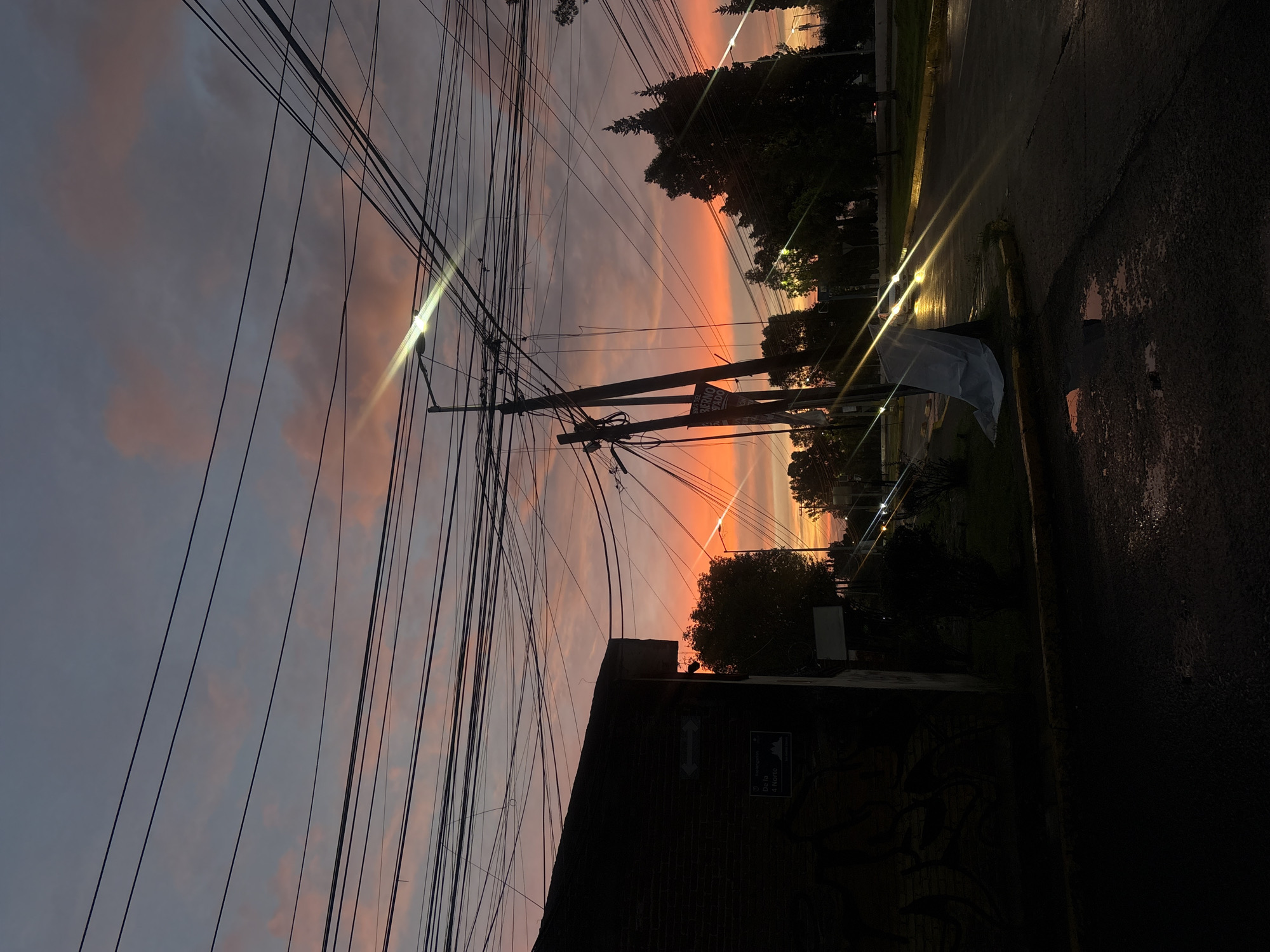 This is a photograph taken at dusk in the city of San Andrés Cholula, Mexico. The sky is painted with vibrant orange and pink hues, with scattered clouds. A dense network of overhead power lines crisscrosses the frame, obscuring parts of the sky and creating a stark contrast against the colorful sunset.
On the left, a dark, brick building with graffiti on its facade occupies the foreground. A street sign is visible on the side of the building. In front of the building, a sidewalk and a street are visible, with puddles reflecting the sky's colors, suggesting recent rain.
To the right, a street curves away, lined with trees and streetlights. A utility pole stands prominently, laden with more electrical wires and supporting a street lamp. The street is wet, and car headlights create streaks of light. A white tarp is draped over a small tree or post near the roadside. The overall scene conveys a sense of urban twilight, with the natural beauty of the sunset juxtaposed against the utilitarian infrastructure of the city.