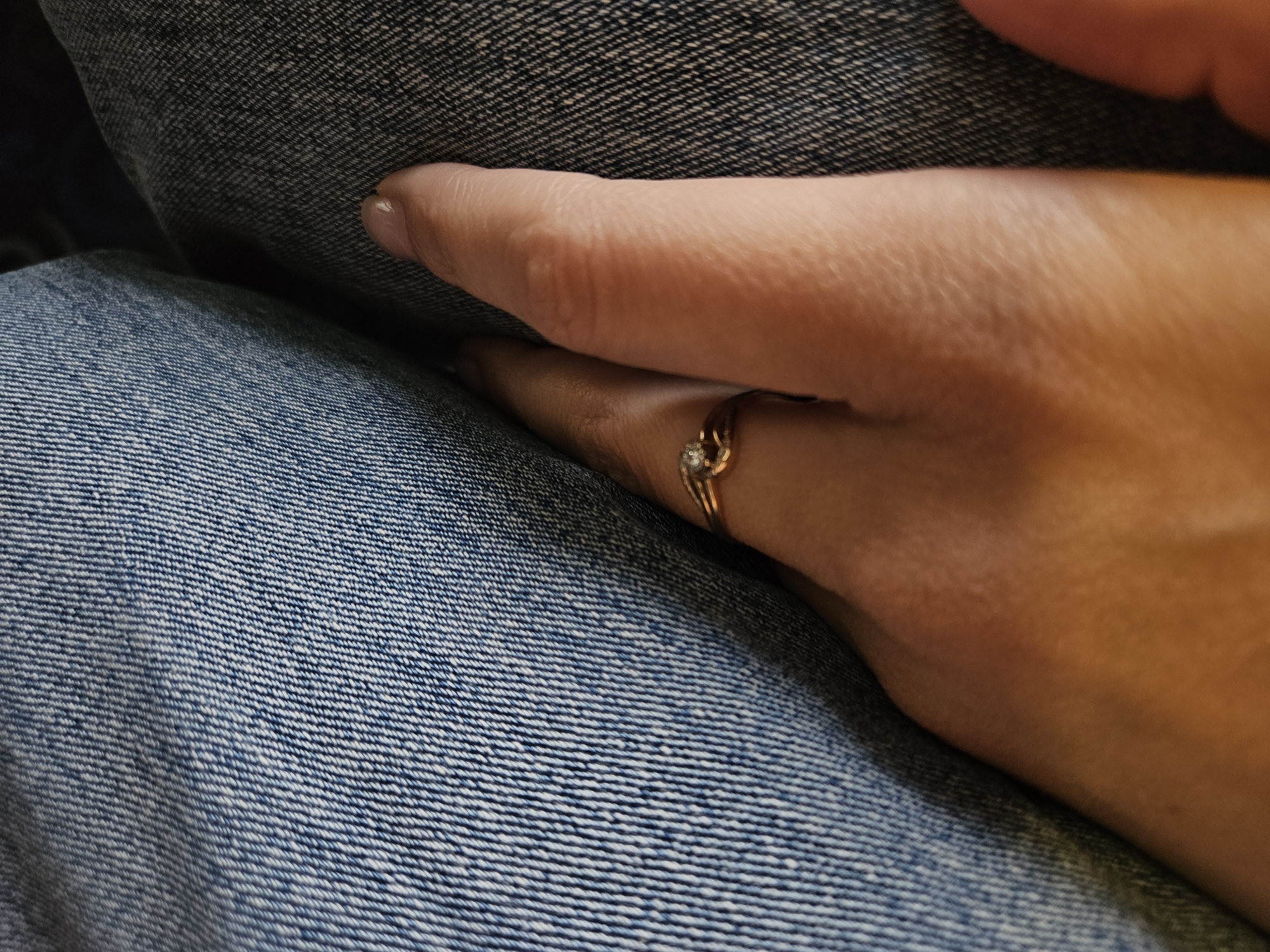 A hand rests on denim-clad legs. The ring finger wears a gold ring with a small stone. The denim has a distinct weave pattern and varying shades of blue. The scene is indoors, illuminated by natural light. There is no indication the image was taken in Chaikovsky, Russia.