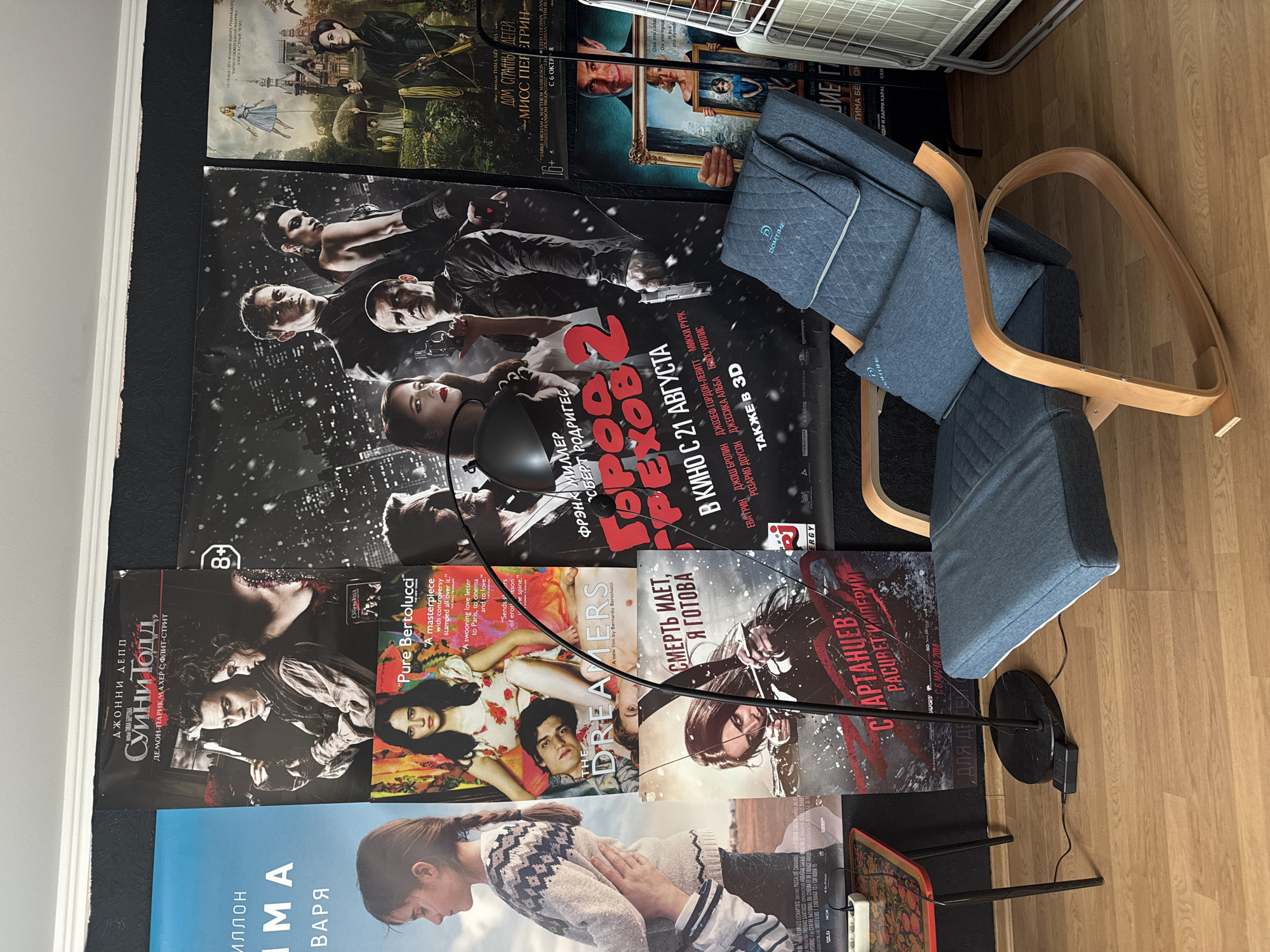 The image captures an indoor room in Krasnodar, Russia, with numerous movie posters adorning a dark-colored wall, displayed horizontally across the frame. The floor is covered with light wood laminate.
A key subject is a light blue/grey upholstered chair with a curved wooden frame, positioned in a reclined state on the right side of the image. A pillow on the chair has the brand name "DOMTIME" visible. Next to the chair is a black modern floor lamp with a spherical head and a circular base, its cord extending towards a power outlet near a small, ornately patterned wooden tray. In the upper right corner, part of a white clothes drying rack is visible.
The wall is heavily decorated with movie posters, primarily featuring Russian titles and promotional text. Visible titles include:
* "Суини Тодд, ДЕМОН-ПАРИКМАХЕР С, ФЛИТ-СТРИТ" (Sweeney Todd: The Demon Barber of Fleet Street) featuring Johnny Depp.
* "THE DREAMERS" by Bernardo Bertolucci, accompanied by critical quotes like "Pure Bertolucci" and "A masterpiece with controversy stamped all over it."
* "СМЕРТЬ ИДЕТ, Я ГОТОВА" (Death Is Coming, I'm Ready), showing a female character with a bow.
* "Город ГРЕХОВ 2" (Sin City 2: A Dame to Kill For), with actors listed including Frank Miller, Robert Rodriguez, Mickey Rourke, Jessica Alba, and Rosario Dawson. It mentions release "В КИНО С 21 АВГУСТА" (In cinemas August 21st) and "ТАКЖЕ В 3D" (Also in 3D).
* "САРТАНЦЕВ: РАСЦВЕТ ИМПЕРИИ" (Sartantsev: Dawn of the Empire), dated "С 6 ИЮНЯ 2013" (From June 6, 2013).
* "ДОМ СТРАННЫХ ДЕТЕЙ МИСС ПЕРЕГРИН" (Miss Peregrine's Home for Peculiar Children), featuring Eva Green, release date "С 6 ОКТЯБРЯ".
* A poster showing a girl, possibly for a film titled "ИЛЛОН ИМА ВАРЯ".
* Another poster with fragmented text "PROTHOMAS SANTUCCI COP..." and "INDEFILMS".
The scene suggests a personalized space, likely a home, belonging to a film enthusiast, given the variety of movie posters acting as wall decor. No specific activity is taking place; it's a static depiction of a room's arrangement. The overall impression is one of a comfortable, albeit cluttered, fan-centric environment.