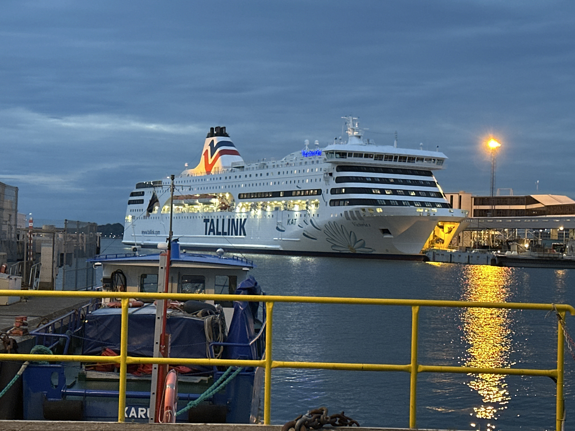 In Tallinn, Estonia, a large white ferry, the "Tallink Victoria I," is docked at a port. The ferry has a distinctive red, white, and blue striped funnel and the "Tallink" logo is prominently displayed on its side. The ferry is illuminated, suggesting either early morning or evening. In the foreground, a smaller blue boat with a blue tarp and yellow railings is moored. The water reflects the lights from the ferry and a nearby lamppost. The sky is overcast with a dusky blue hue. The scene appears to be a typical port environment with industrial structures and the calm water of a harbor. There are no people visible in the image.