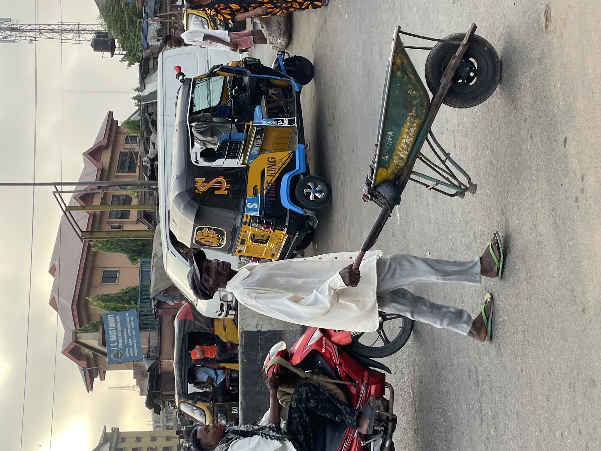 This image captures a bustling street scene in Onitsha, Nigeria, under an overcast sky, suggesting either early morning, late afternoon, or a cloudy day. The setting features a paved road with various modes of transport and buildings lining the street.
In the foreground, a man in a white long-sleeved shirt and grey trousers, wearing green flip-flops, pushes a green and yellow wheelbarrow. His gaze is directed towards the left side of the frame. To his right, another person is seated on a red motorcycle, which has "Hoalite" visible on its side.
Behind the wheelbarrow man, a prominent yellow and black tricycle, commonly known as a Keke Napep, is visible. It features blue accents and displays text including "FUTURE KING" and an "S" logo. Further back, a white van is partially visible. Several other people are scattered throughout the scene, some partially obscured by vehicles or buildings, including an individual wearing patterned clothing near the white van.
A blue signboard for "JC MASS TRANSIT" stands by the roadside, listing destinations such as "EZEIWEKA BUS TERMINAL OBOSI", "ONITSHA", "PORTHARCOURT", "ONITSHA-ELENE, CYIGBO ELENE JUNCTION", and "RUMUONDRO MILE 3-MILE 1 MATER LINE," confirming the Nigerian location. In the background, residential or commercial buildings with red roofs are visible, along with a tall telecommunications tower and a water tank on a stand. The scene depicts a typical day of movement and commerce in an urban Nigerian environment.
