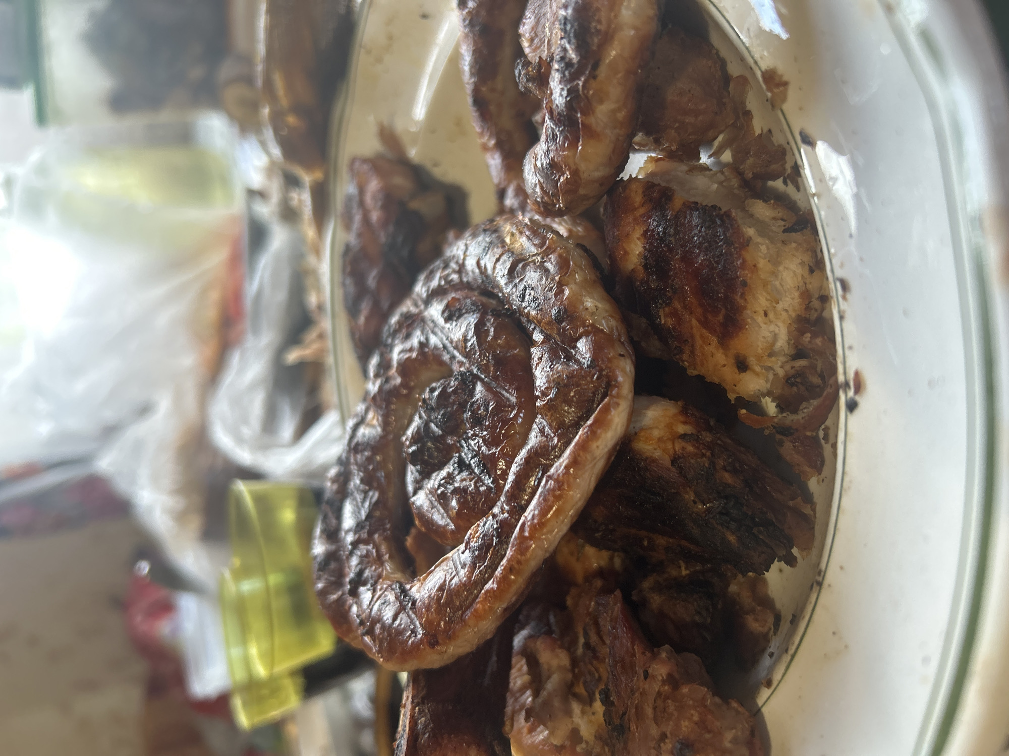 A close-up shot depicts food in a transparent container. The food includes a coiled sausage-like pastry, browned, and what appears to be cut pieces of cooked meat, possibly chicken, also browned. The container's edge is visible. The surrounding environment is blurry and indistinct. There is a suggestion of other objects, including what may be a container with a yellowish label. The location is unspecified within the image, and there is no forensic evidence related to the city of Chaykovsky, Russia.