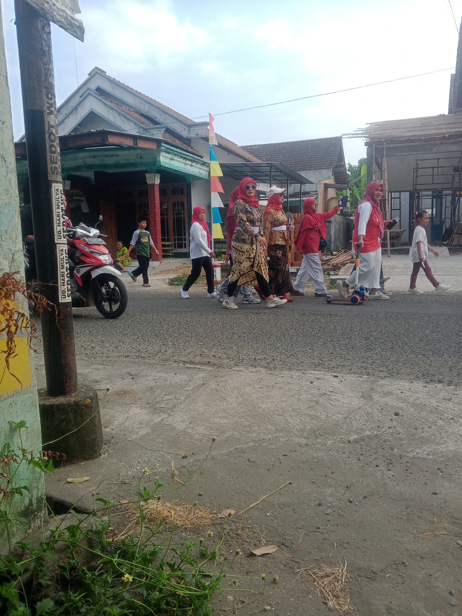 In Lembah, Indonesia, a group of individuals, predominantly women wearing headscarves and red or patterned clothing, are walking on a paved road. The group includes at least six adult women and one young girl. One woman in the foreground is holding a phone and appears to be recording. To their left, a red and white motorcycle is parked. In the background, houses with visible roofs and construction scaffolding are present. A boy in a green shirt is walking away from the motorcycle. A colorful string of flags hangs across the street in the middle distance. The road surface is composed of asphalt and concrete. Greenery, including grass and plants, borders the road. A utility pole stands on the left side of the image, with visible stickers and text. The sky is overcast.