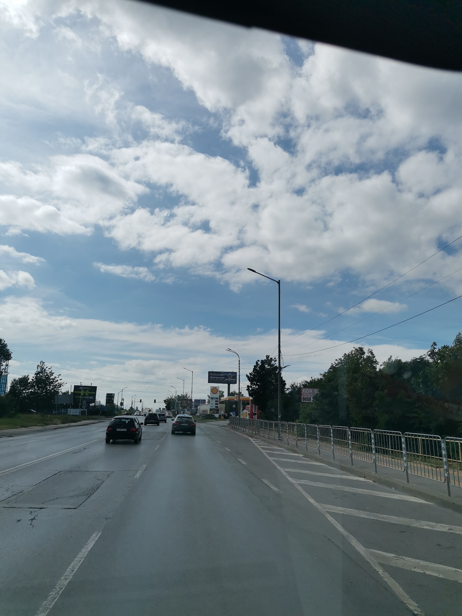 The image depicts a daytime scene on a wide, wet asphalt road in Sofia, Bulgaria. The road has multiple lanes and is bordered by trees and roadside structures, including billboards and streetlights. Several vehicles are visible on the road, including cars and what appears to be a van or small truck. The sky is partly cloudy with a mix of blue sky and white cumulus clouds. The overall atmosphere suggests a suburban or commercial area on a possibly overcast or recently rained-on day.