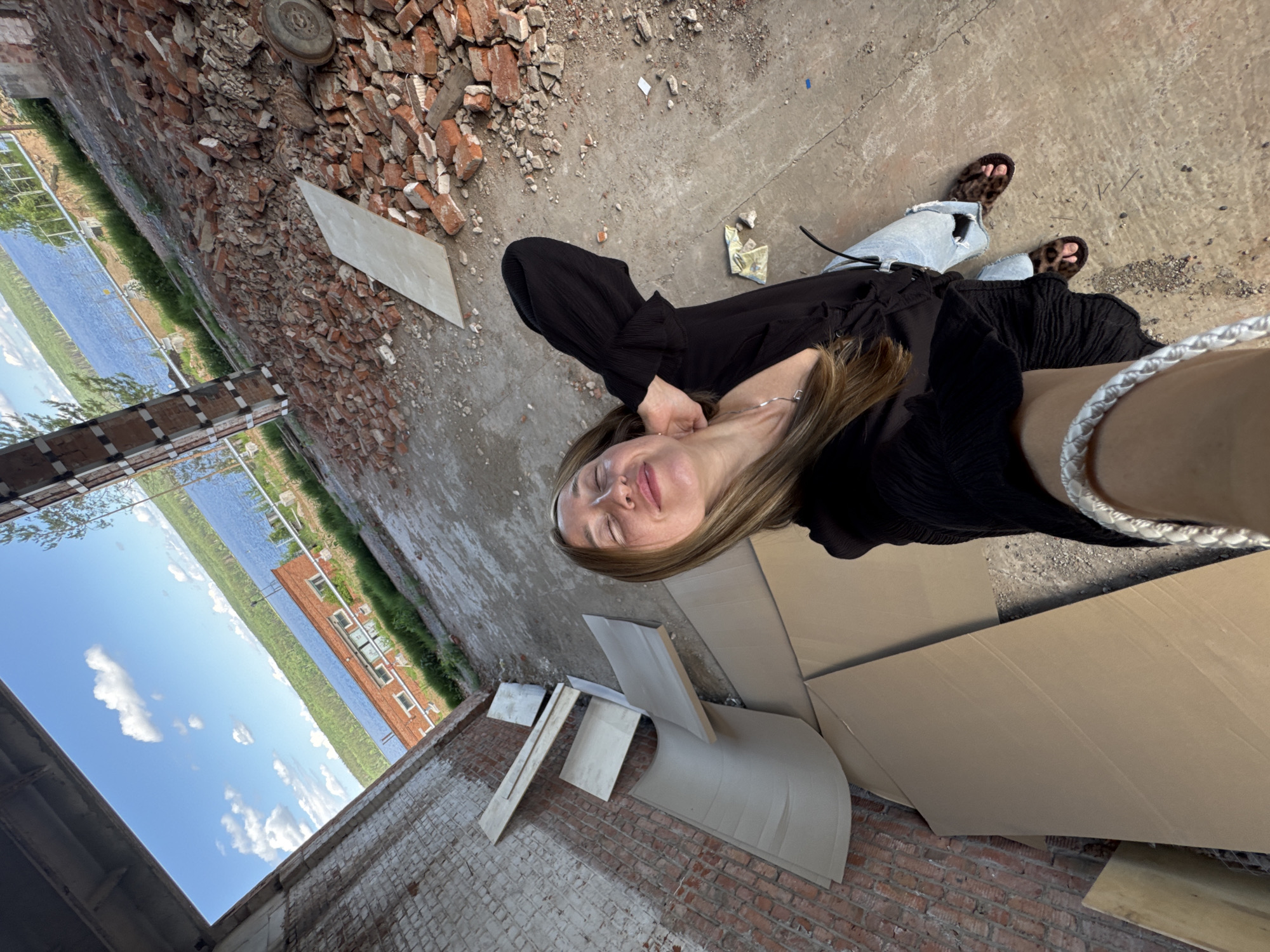 The image depicts a woman taking a selfie in an unfinished building. The environment is characterized by exposed brick walls, concrete flooring, and construction debris, including piles of broken bricks and large cardboard sheets. The woman is wearing a black top, light blue jeans, and leopard-print sandals. She has blonde hair and is smiling, with her eyes closed. A silver necklace is visible around her neck, and a white braided bracelet is on her left wrist.
The background reveals a body of water, possibly a river or lake, with a forested shoreline and a brick building visible across the water. The sky is blue with white clouds. The scene is likely located at an industrial or construction site near a body of water. The context provided by the user suggests this location is in the city of Chaikovsky, Russia, specifically near the Kama River, which is a prominent geographical feature of the area. The unfinished state of the building, combined with the natural landscape in the background, suggests an area undergoing development or renovation.