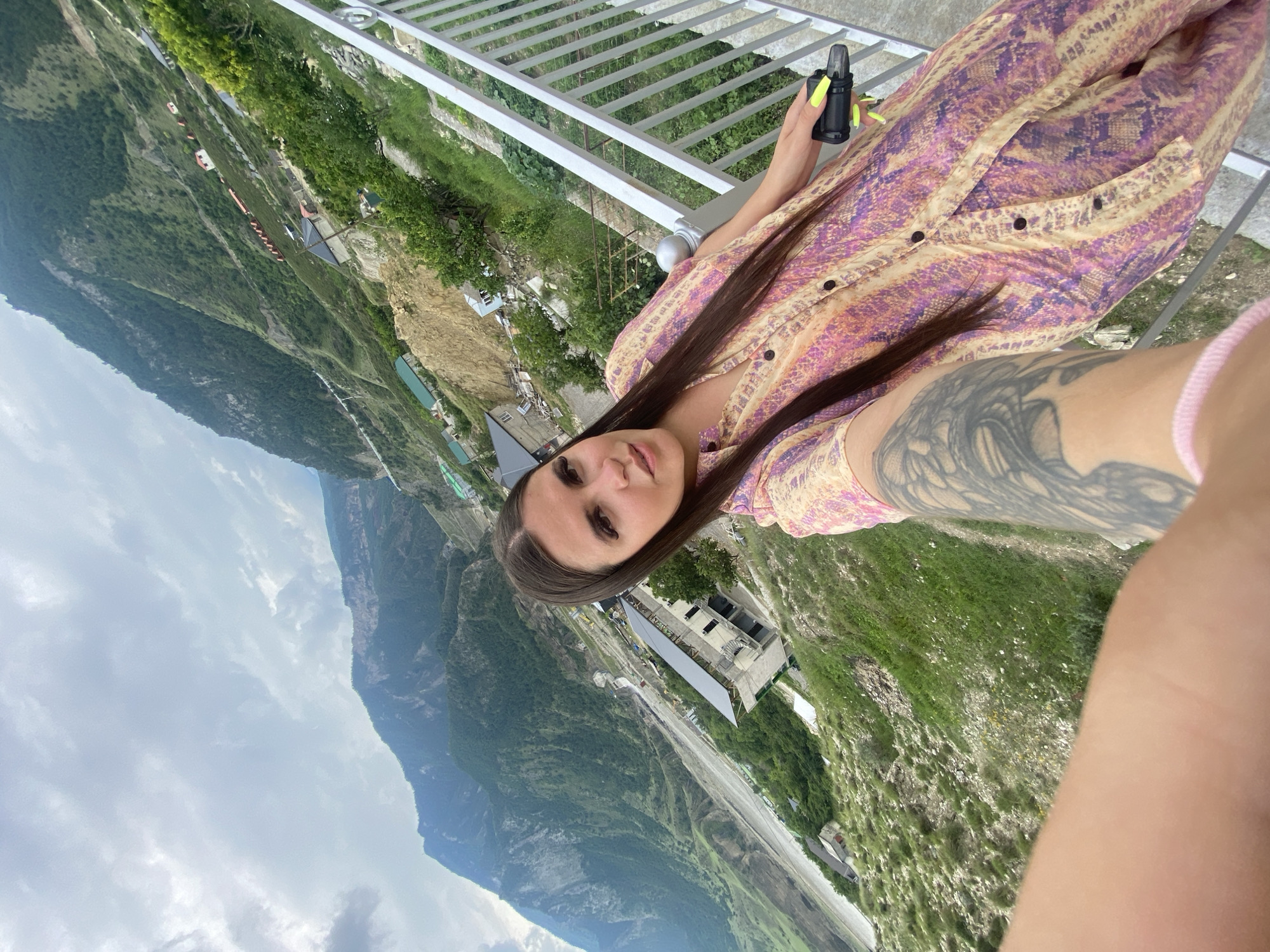 A woman with long, dark hair and a patterned dress takes a selfie on a balcony overlooking a mountainous landscape. The woman is holding a black object with a neon green tip in her right hand. The mountainous terrain features green vegetation, scattered buildings, and a landslide. The sky is cloudy. The location appears to be Chalda, Russia, based on the prompt.