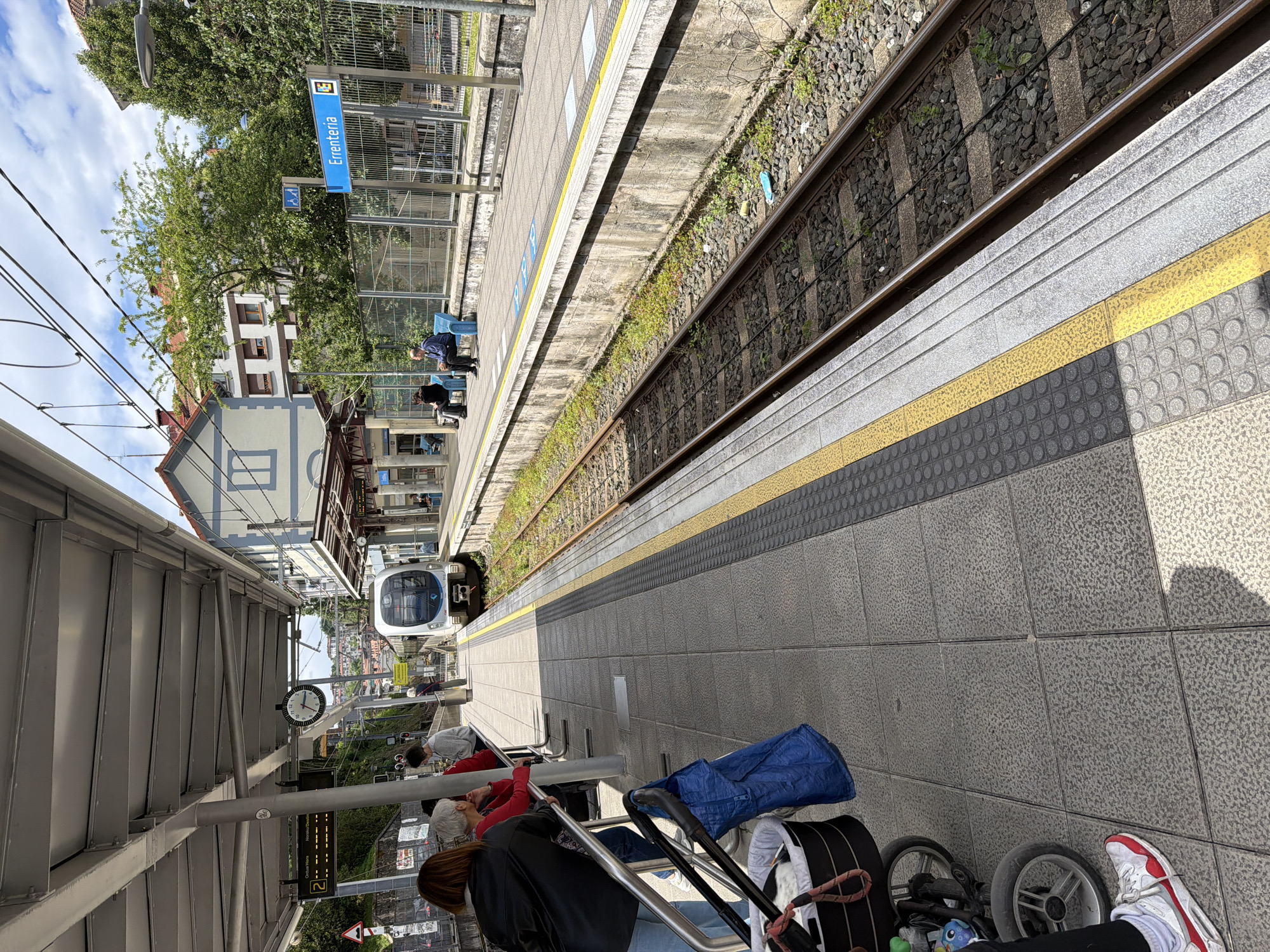 This image shows a train station platform in Errenteria, Spain. The platform is lined with gray tiled flooring and has a yellow safety line along the edge. A train is approaching the station on the tracks next to the platform. Several people are waiting on the platform, including some seated on benches. A station sign with the name "Errenteria" is visible. Overhead, the station has a metal roof structure and streetlights are visible in the background.