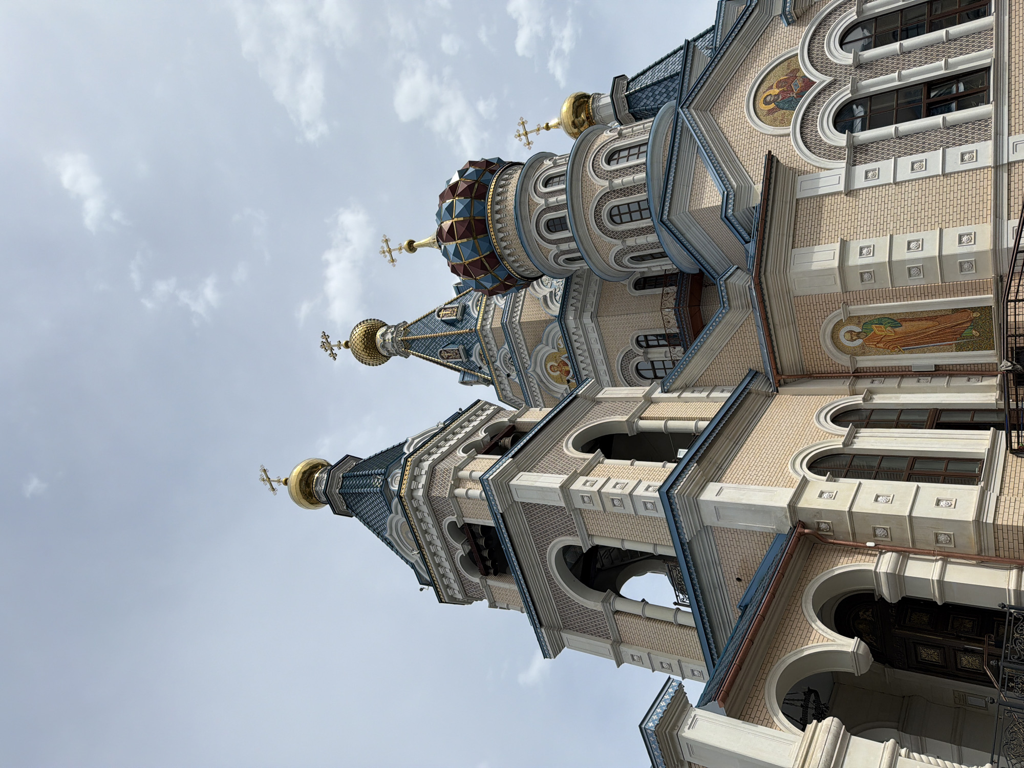 The image showcases the architecture of St. Michael's Cathedral in Izhevsk, Russia. The view is a low-angle perspective, emphasizing the building's height and intricate detailing. The cathedral features multiple towers topped with golden domes and crosses. The walls are constructed with light-colored brick, embellished with arched windows, columns, and decorative mosaic panels. The sky is overcast, providing a neutral backdrop. The overall impression is one of grandeur and ornate religious architecture.