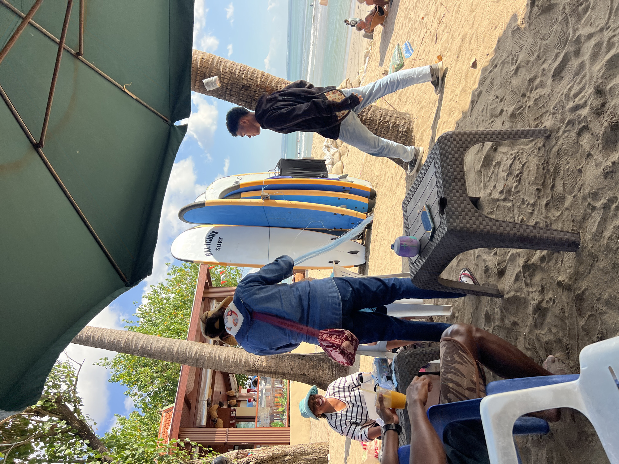 In Kuta, Indonesia, on a sandy beach under a large green umbrella, several people are gathered. A woman wearing a blue denim jacket and jeans stands near a rack of surfboards. She has a patterned bag slung across her shoulder. To her left, a woman in a striped shirt and a blue hat sits at a table with a glass of orange juice. In the background, a young man in a black hoodie and light blue jeans stands near a palm tree, looking out towards the ocean. The beach has several tables and chairs, and the ocean is visible with gentle waves. There are palm trees and some buildings in the distance, suggesting a commercial area near the beach. The overall scene depicts a relaxed atmosphere on a sunny day.