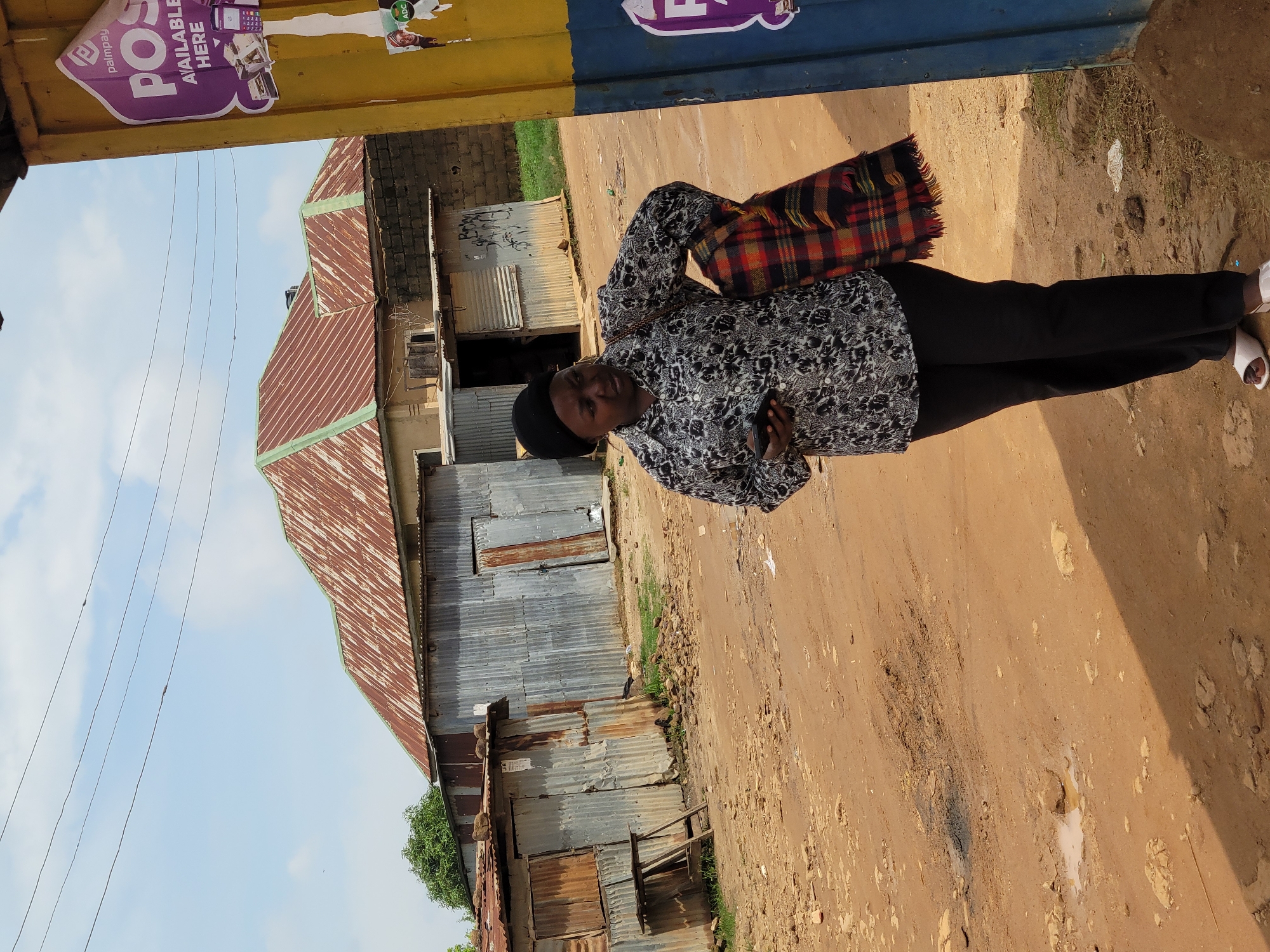 A woman wearing a black headwrap, a black and white patterned shirt, and black pants stands on a dirt road in Jos, Nigeria. She is holding a smartphone in her right hand and has a red, orange, and black plaid scarf draped over her left shoulder. Behind her, there are several corrugated metal structures, some with rusted roofs. A yellow and blue pole with advertisements is to her right. The sky is partly cloudy.