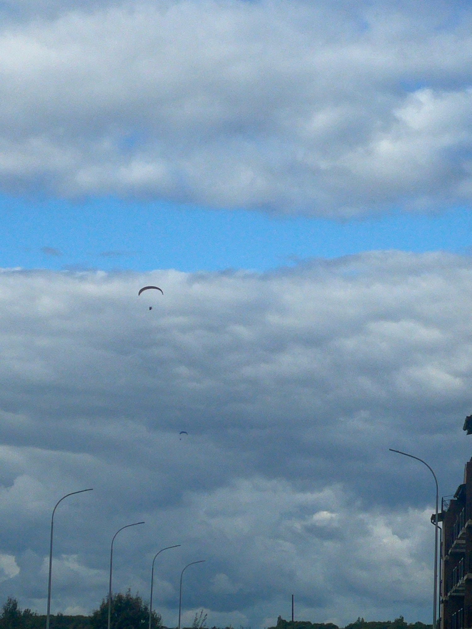 A paraglider is visible in the sky above Kornevo, Russia, soaring amidst a cloudy atmosphere. The paraglider is small and dark against the backdrop of a bright blue sky segment bordered by thick, grey clouds. Below the paraglider, a light pole with a curved arm stands tall, and other similar light poles are lined up along what appears to be a road or pathway. A portion of a building with balconies is visible on the far right, indicating an urban or suburban setting. The overall scene depicts a moment of recreational activity in the sky over a developed area.