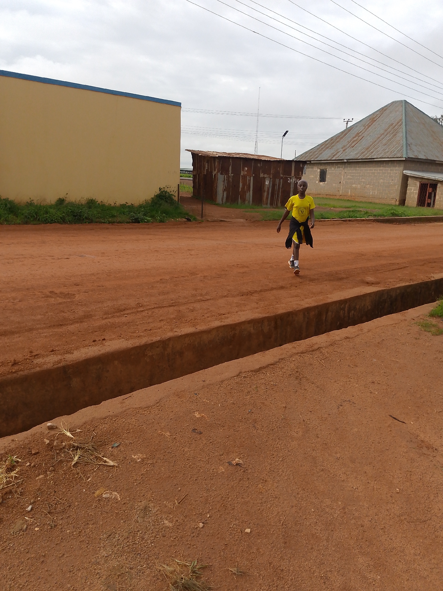 In Barakin Angundi, Nigeria, under an overcast sky, a person wearing a yellow t-shirt and black shorts walks along a dirt road. The person's t-shirt has a logo on the left chest. A black jacket or sweater is tied around their waist. They are wearing white and orange athletic shoes. The road surface is reddish-brown dirt with visible tire tracks. To the left of the road, there is a beige building with a blue trim along the roofline, bordered by green vegetation. To the right of the road, a corrugated metal structure stands, followed by a building constructed of light-colored bricks with a grey, pitched metal roof. A concrete drainage ditch runs parallel to the road in the foreground. Electrical wires are visible against the sky in the background.