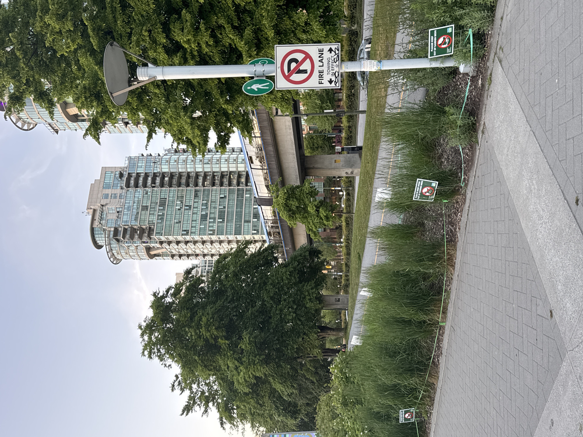 The image shows a streetscape in Vancouver, Canada. In the background is a tall, modern residential building with multiple balconies. An elevated walkway or bridge is also visible. In the foreground is a grey brick pathway, bordered by a grassy strip. Several small signs reading "Please keep out of Garden Beds" are placed along the grassy edge. A white signpost has several signs affixed to it: a pedestrian sign, a bicycle sign, and a "Fire Lane No Stopping Towing in Effect" sign. The scene is bathed in sunlight, indicating a clear day.