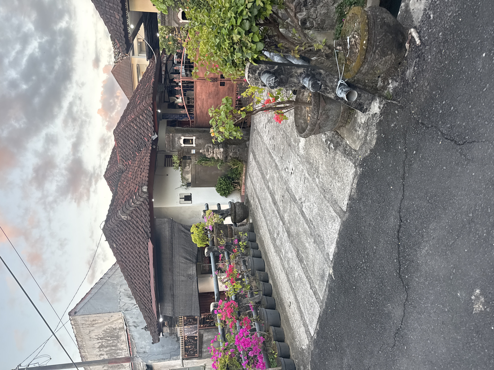 The image depicts a street scene in Denpasar, Indonesia, showcasing Balinese architecture and landscaping. A row of potted bougainvillea flowers lines the walkway, adding vibrant color. A traditional Balinese house, characterized by its red-tiled roof and ornate gate, stands behind the flowers. The sky is overcast with some visible pinkish hues from the sun. The pavement is asphalt with a visible crack running through it. The location seems to be a residential area.