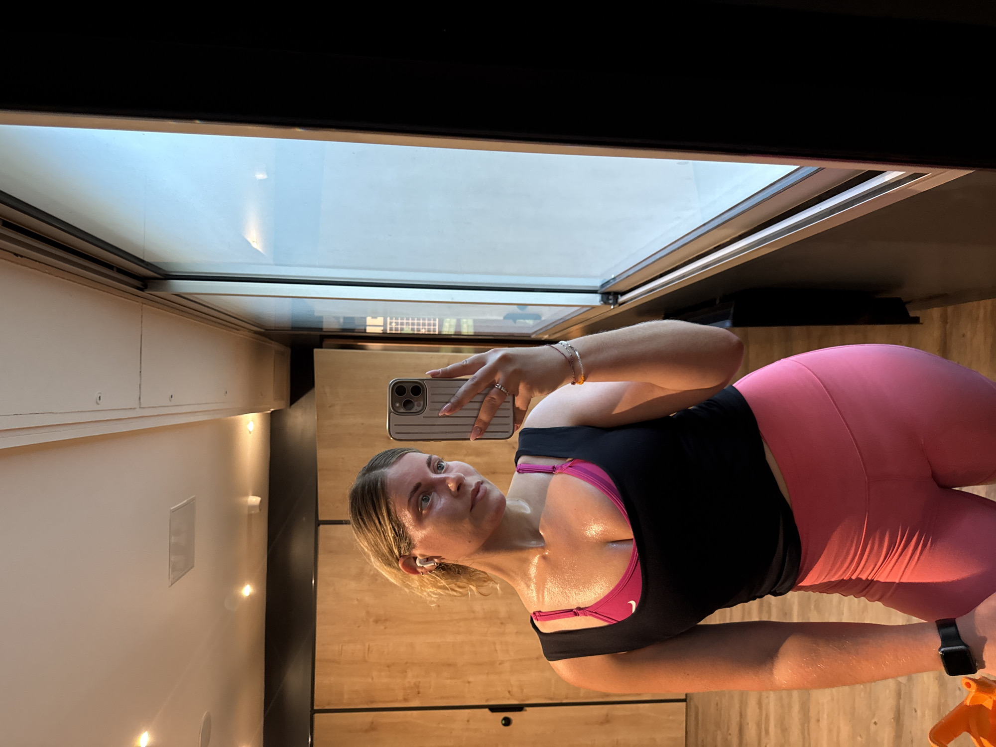 A woman is taking a selfie in what appears to be a gym or fitness studio with wooden walls and flooring. She is wearing a black sports top over a pink sports bra and pink leggings. The woman is sweaty, suggesting she has been exercising. She is holding a smartphone up to take the picture. A window is visible in the background with a view that could potentially be the sky. However, without other landmarks visible through the window, it is impossible to confirm the location is Paris. The gym or studio appears to be indoors and well-lit.