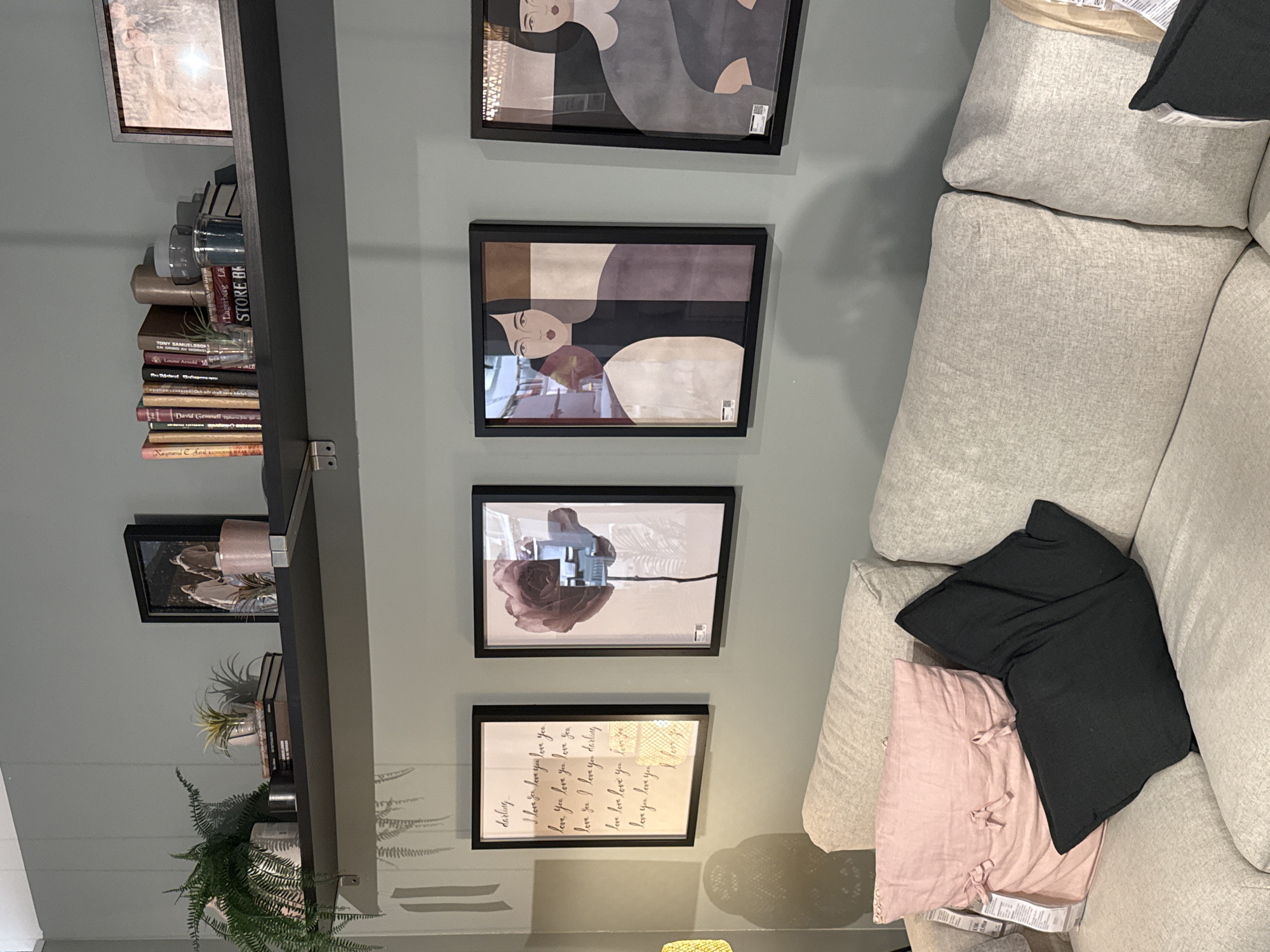 The image shows a shelf against a wall, adorned with books, decorative jars, and plants. Four framed pictures hang on the wall beside the shelf. A sofa is positioned adjacent to the wall, with a black and a pink pillow placed on it. The text in one of the frames is partially readable, mentioning "I love you" repeatedly. Some book titles on the shelf include "TONY SAMUELSSON," "Louise Arnold," "David Gemmell," "Raymond E Send KUNG," "Lagerberg La," and "STORE BE." The image does not contain enough information to identify the location of the scene in Sofia, Bulgaria.